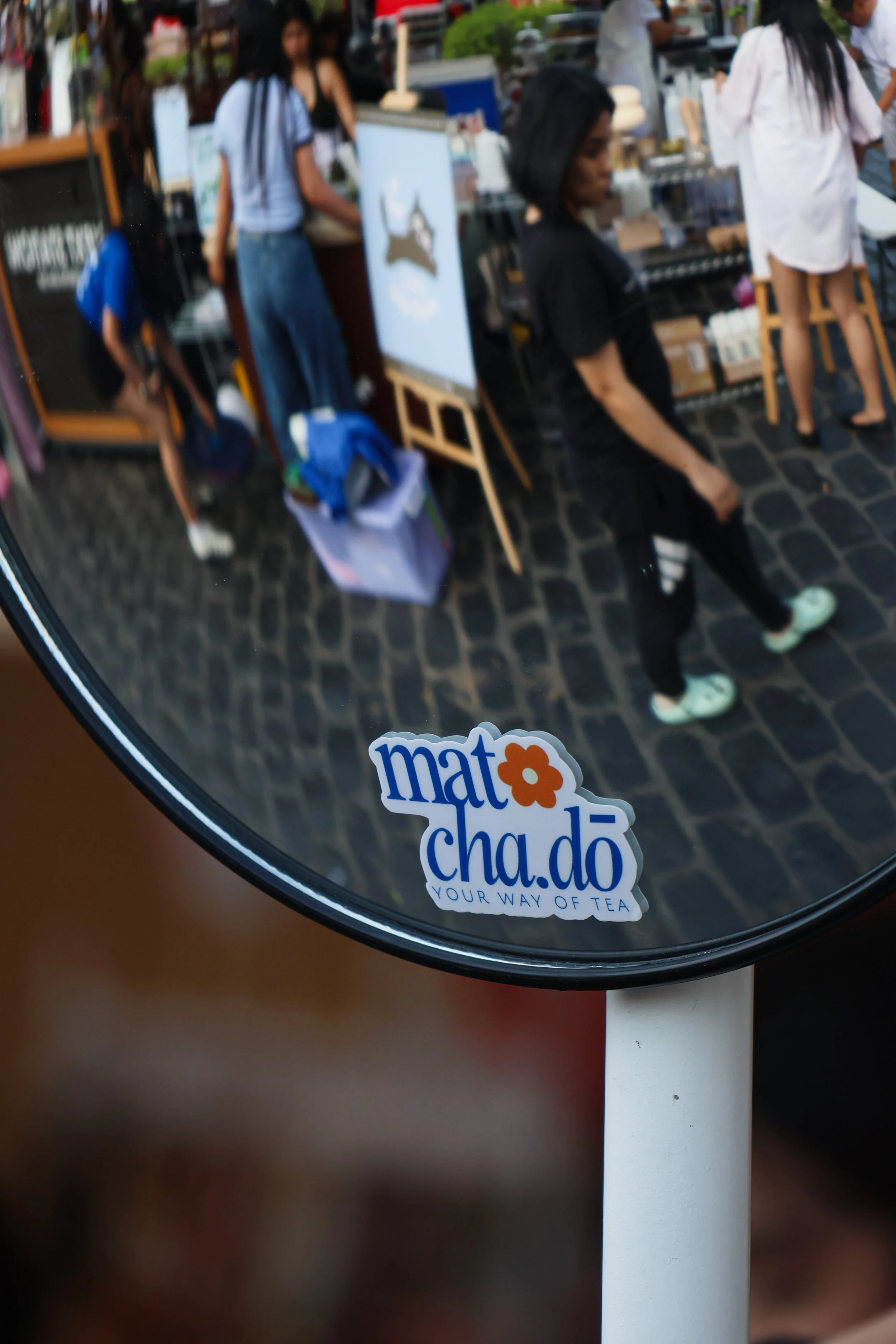 Reflection of people behind a tea stand in a mirror with a sticker that says 'mat cha do' and 'YOUR WAY OF TEA'.