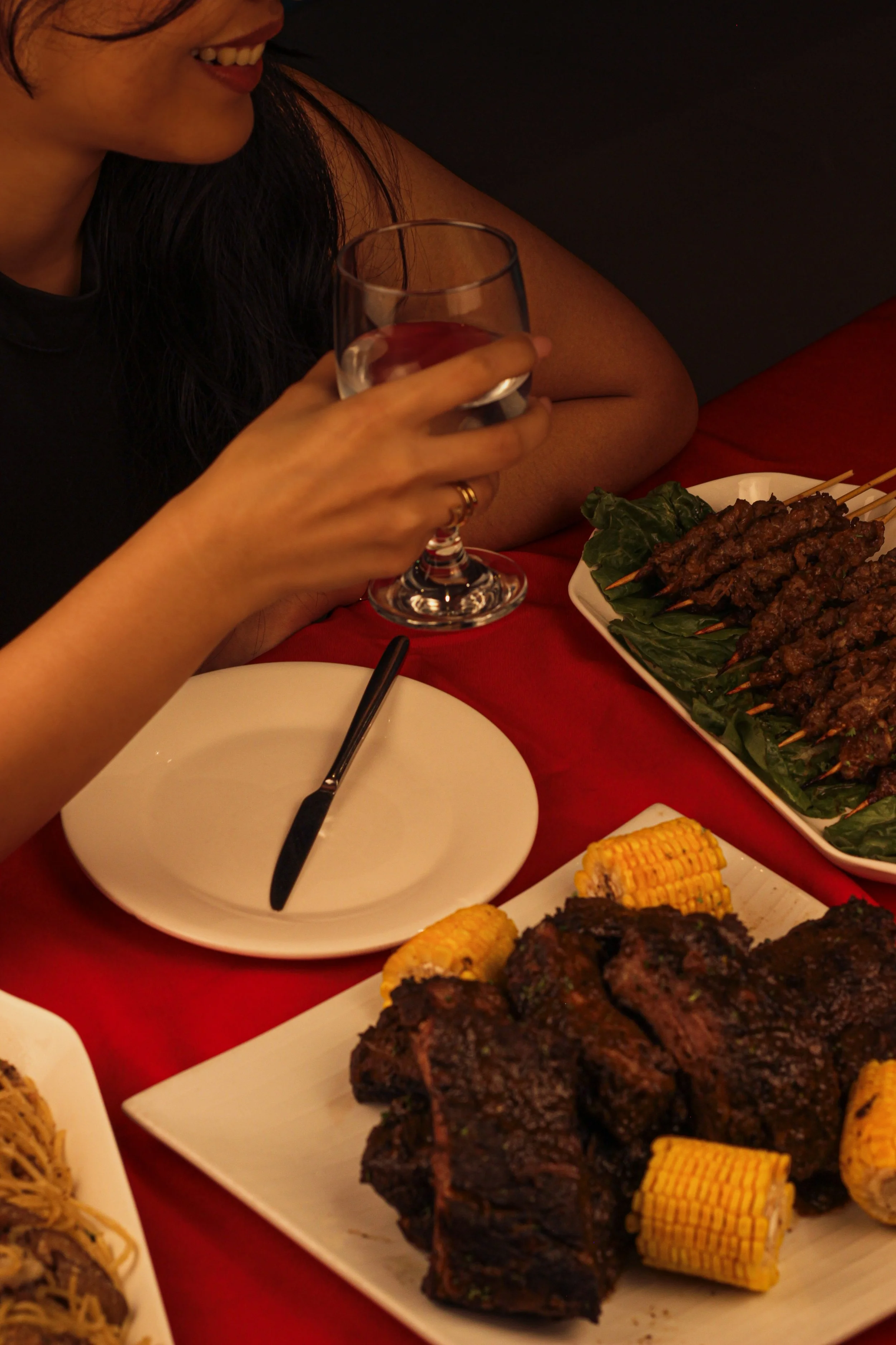 A woman smiling and holding a glass of water at a dinner table with various foods including grilled meat, corn on the cob, and skewers on a red tablecloth.