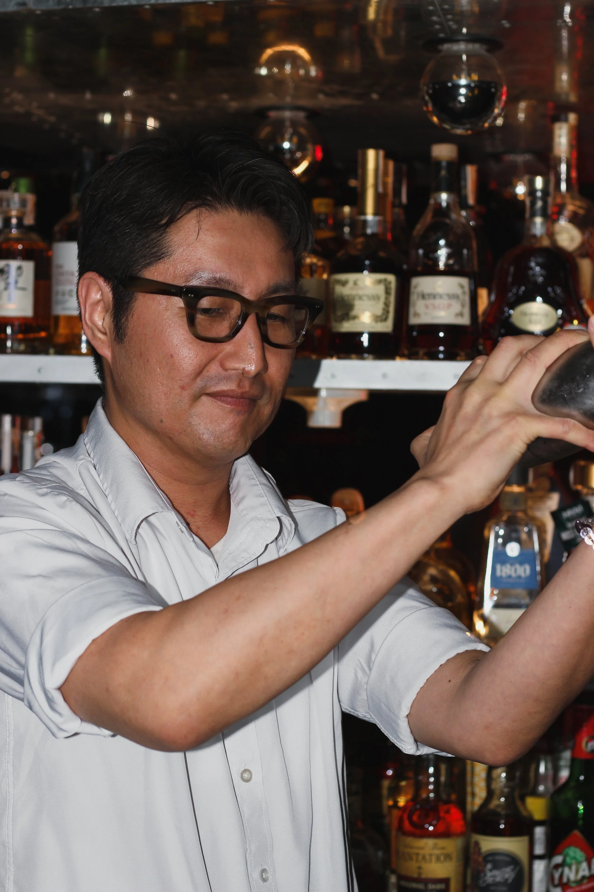 A man with black hair, glasses, and a white shirt is standing behind a bar shelf filled with bottles of alcohol, including whiskey and other spirits.