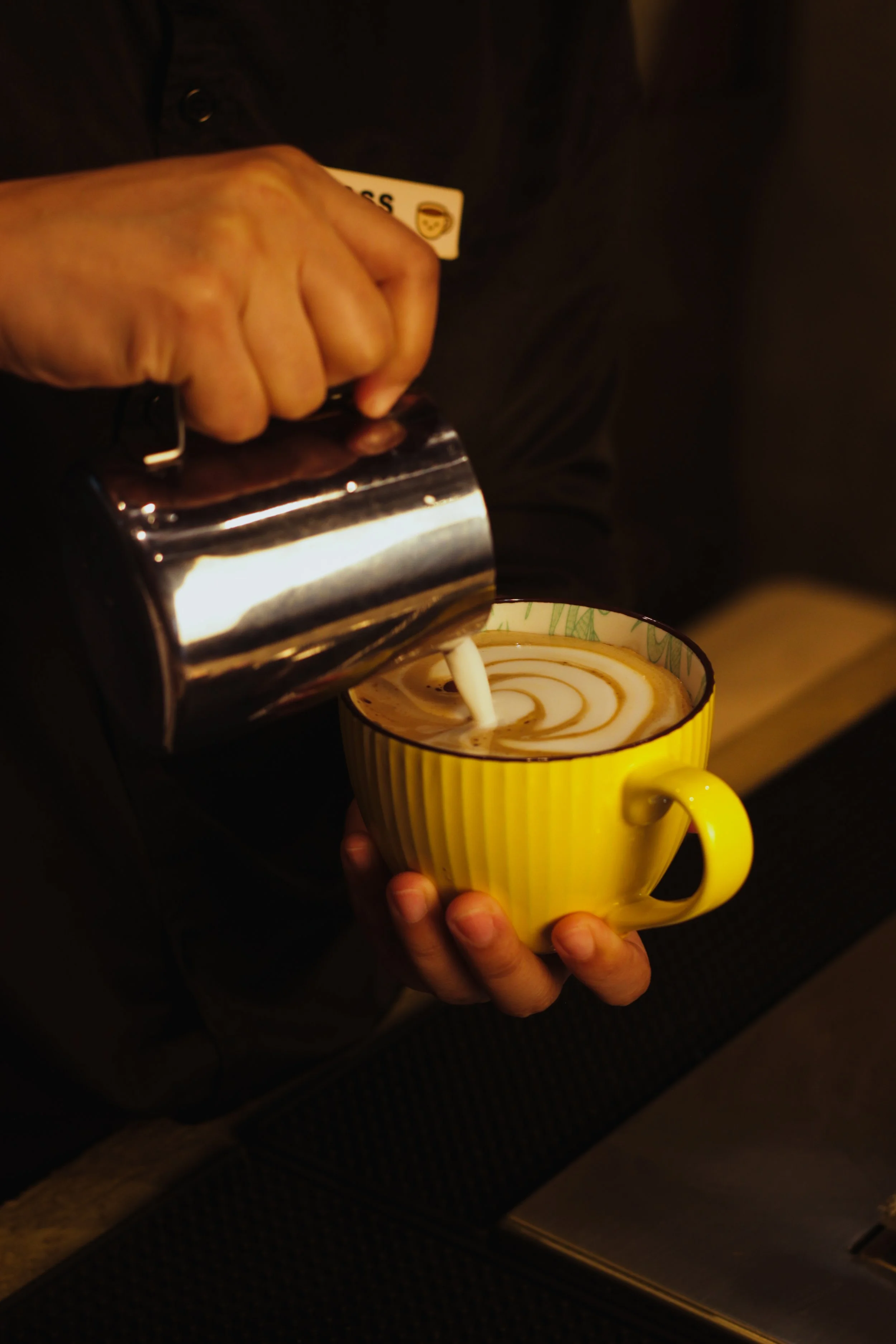 A person pours cream into a yellow mug of coffee with latte art.