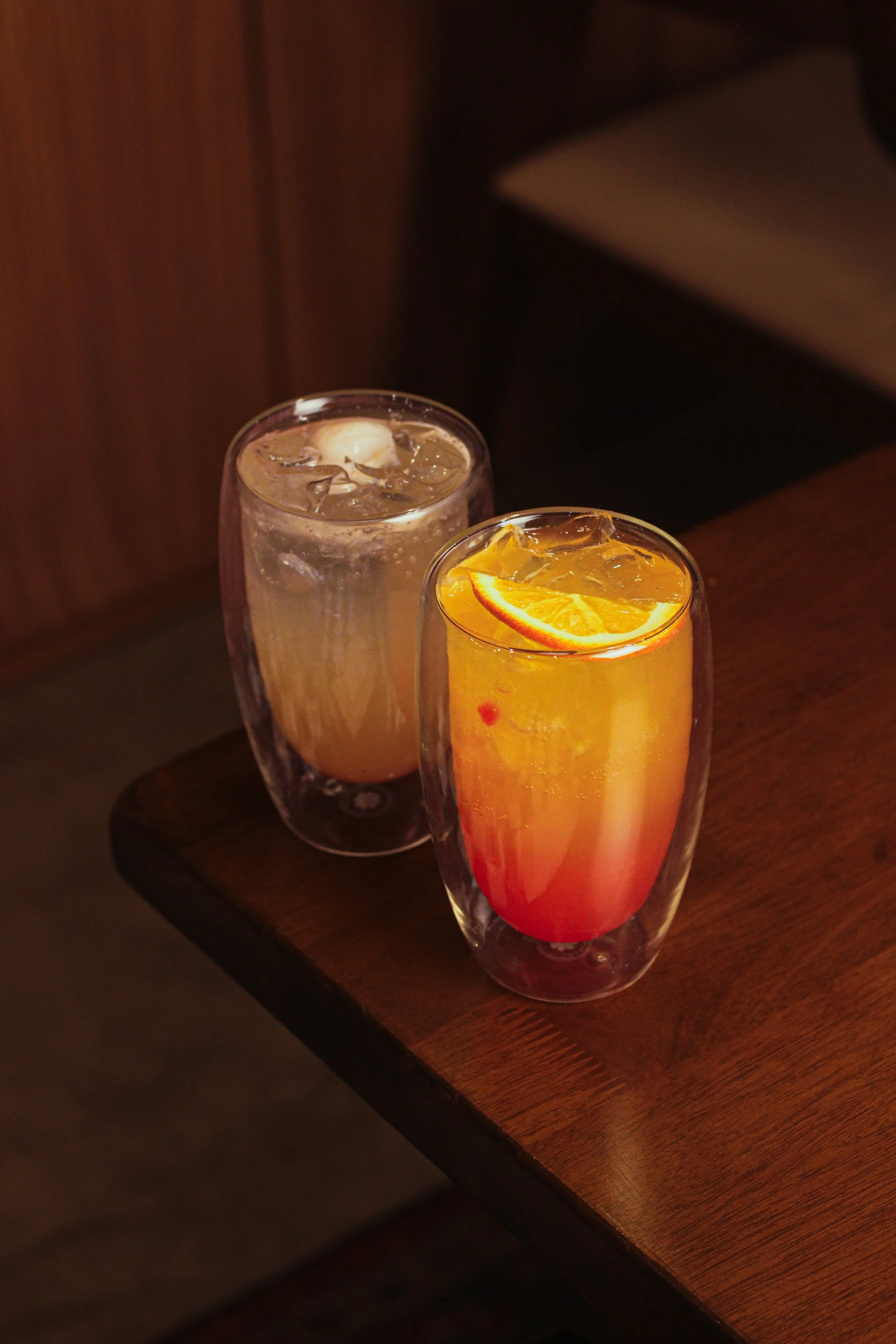 Two colorful beverages in tall glasses on a wooden table, one with an orange slice and the other with ice and a lemon wedge.