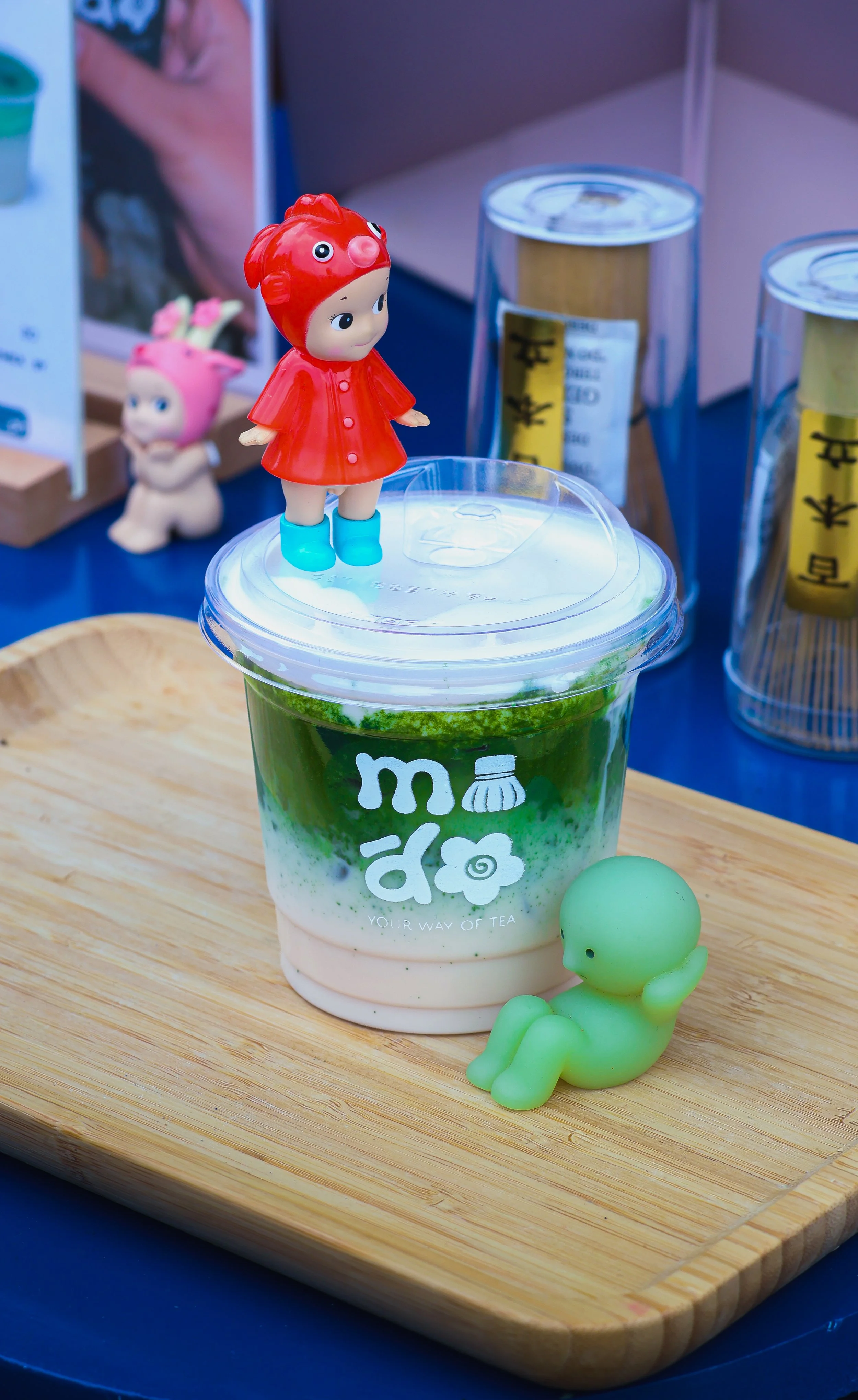 A plastic toy girl wearing a red dress and blue boots standing on a clear plastic lid atop a layered green and white drink in a transparent cup with the words "your way of tea". Next to the drink is a small green rubber duck.