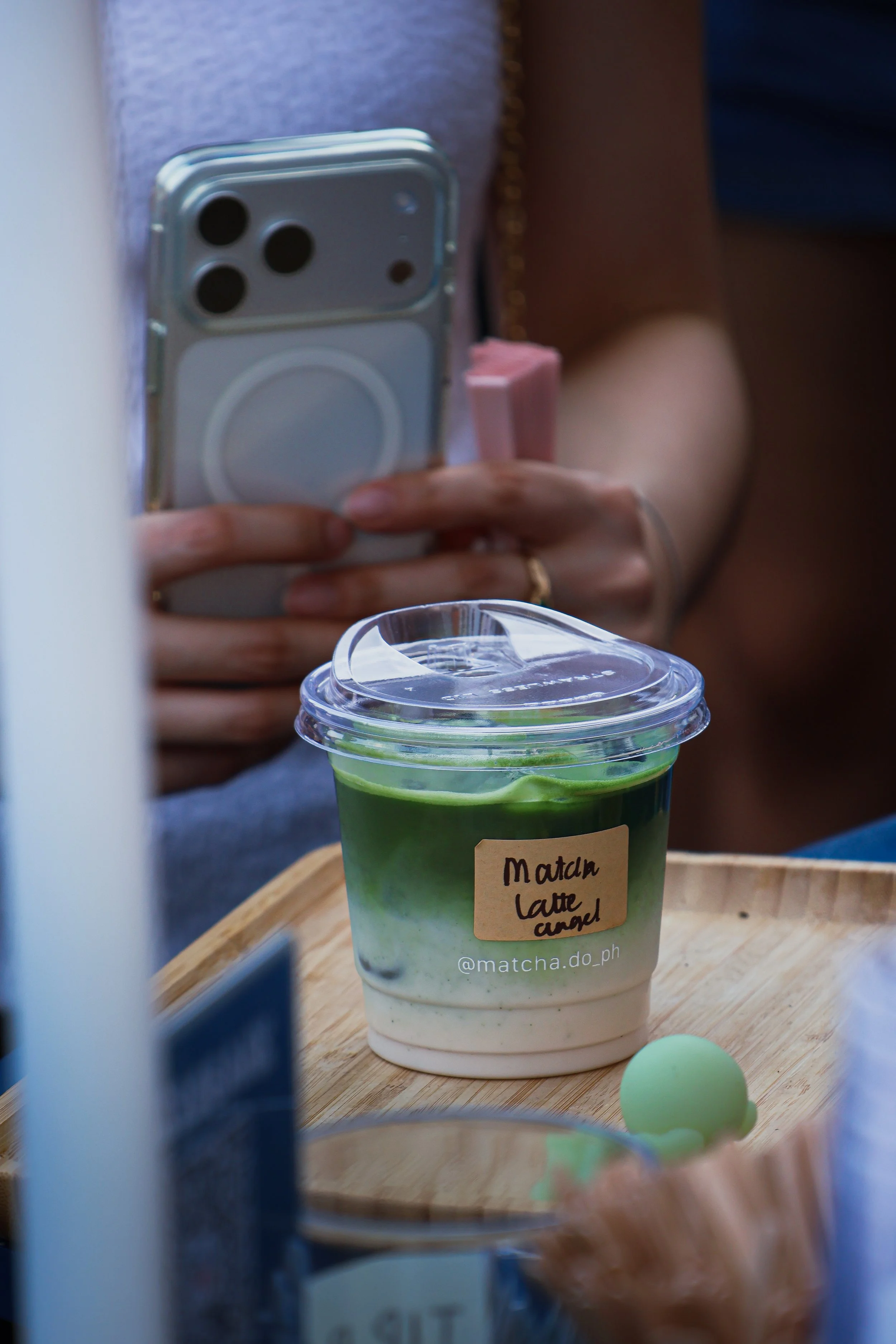 A person holding a smartphone, taking a photo of a matcha latte drink in a clear cup with a green and white layered design, labeled 'Matcha Latte' with a straw, on a wooden tray. There is a cup lid and a pastel green sphere in the foreground.