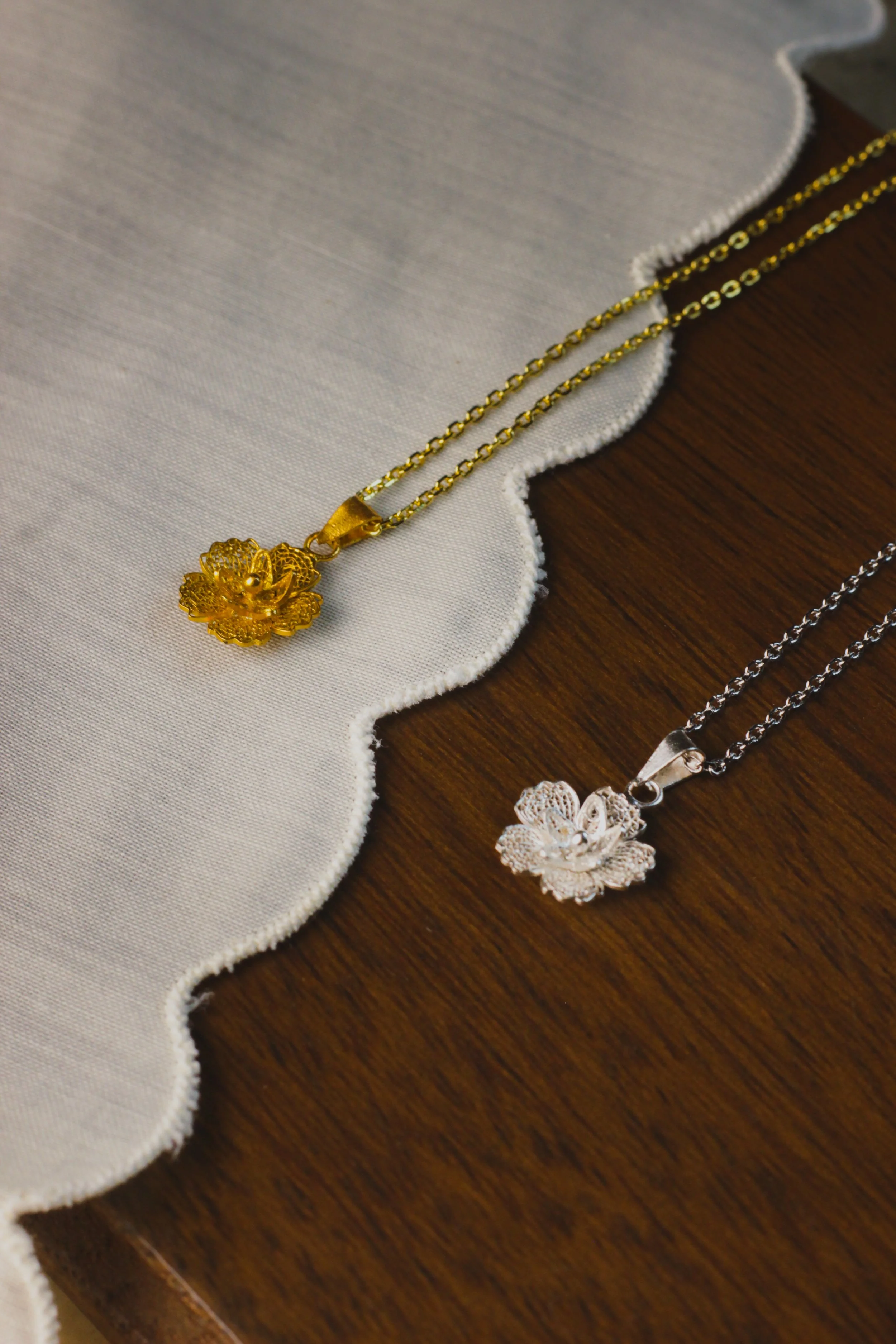 Gold necklace with a floral pendant on a white cloth, and a silver necklace with a pendant on a wooden surface.