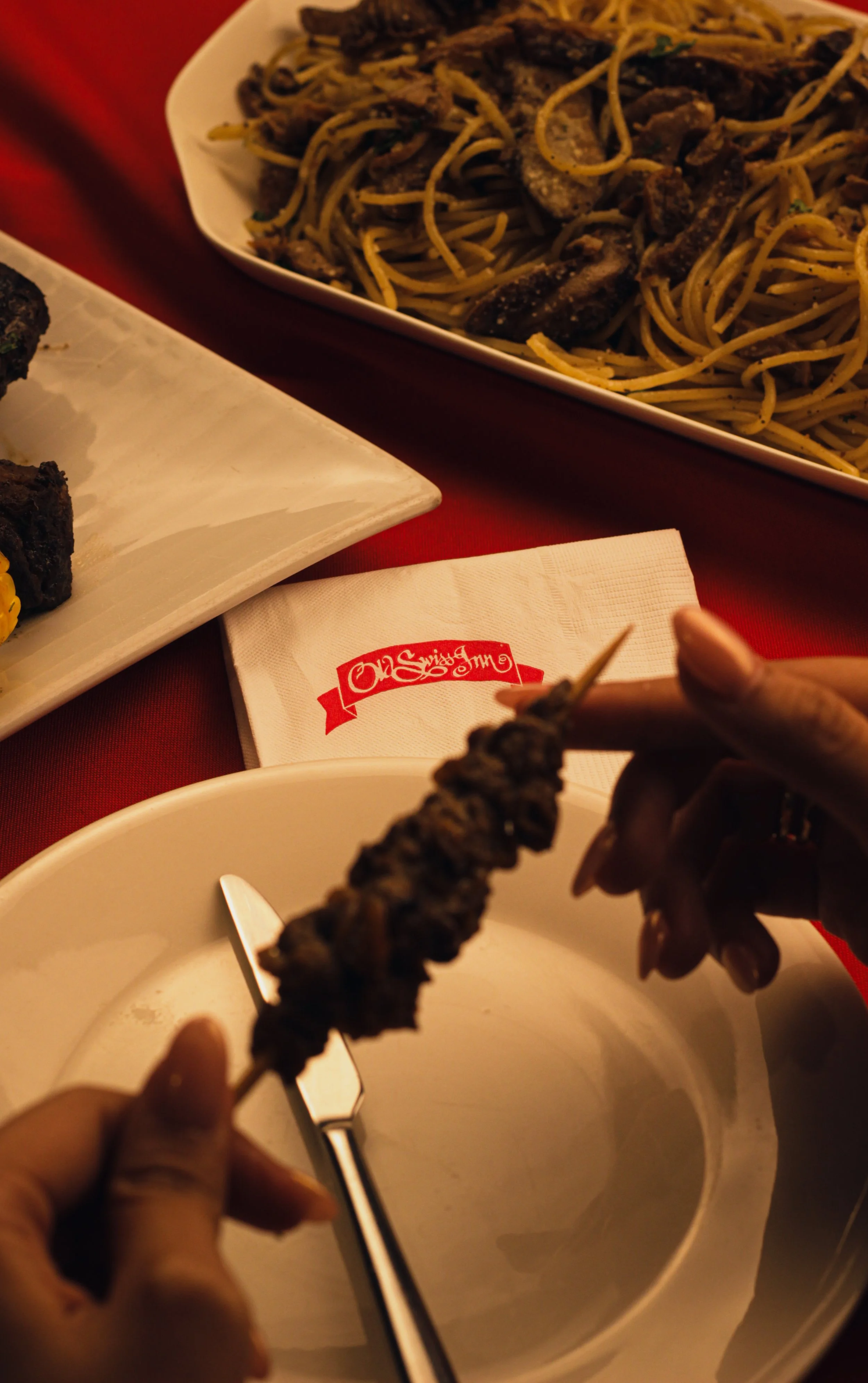 Person holding a skewer of grilled meat over a white bowl, with a plate of cooked pasta in the background and a red tablecloth underneath.