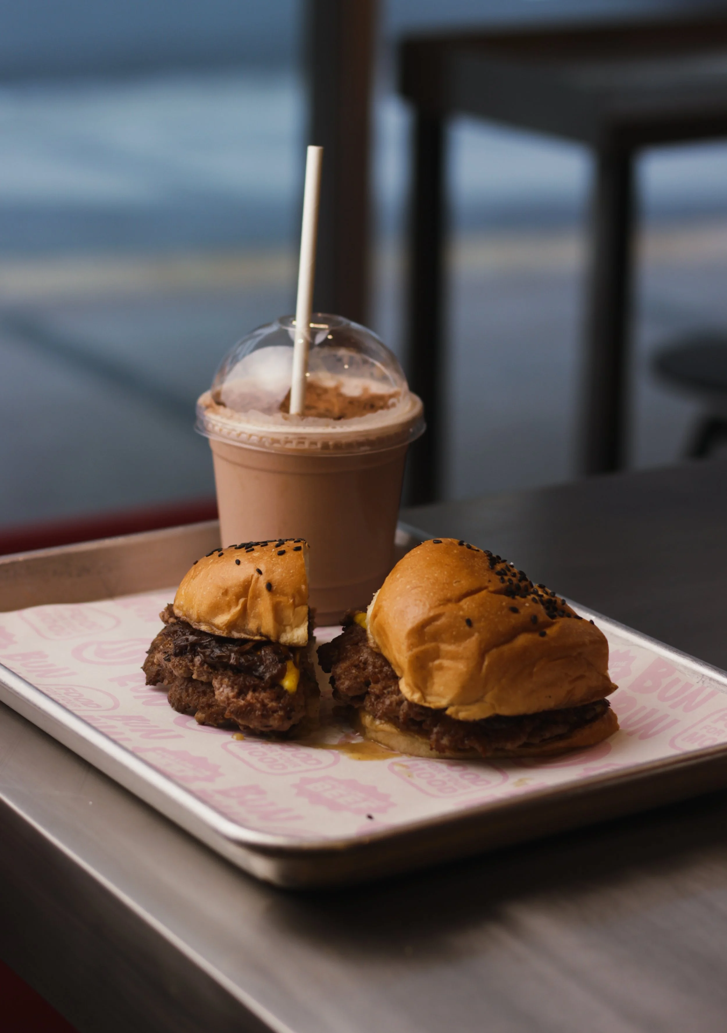 Two cheeseburgers and a milkshake on a tray at a restaurant, with a blurred background of a window and furniture.