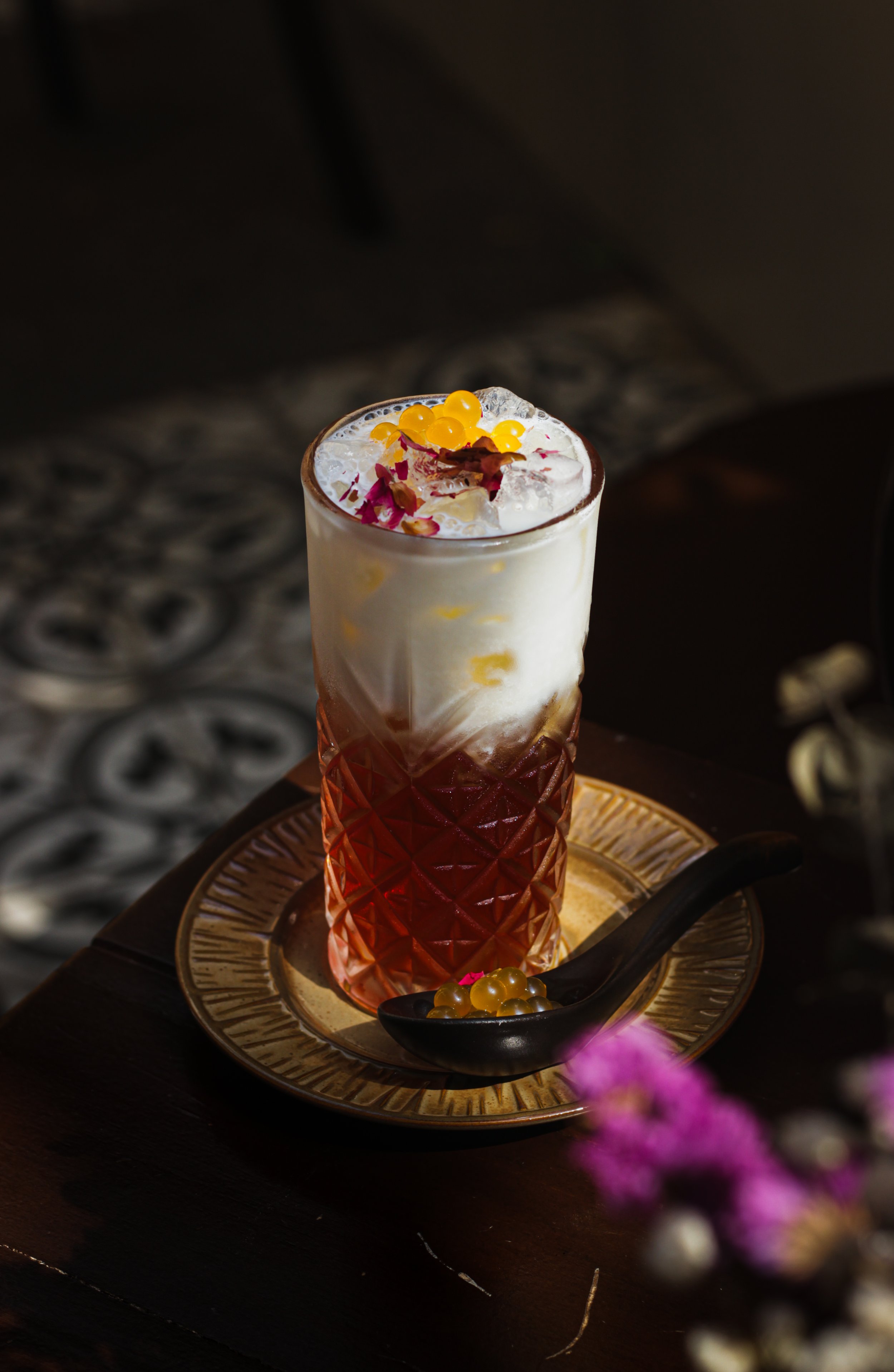 A tall glass of layered drink with whipped cream, yellow tapioca pearls, and pink and red flower petals on top, placed on a decorative gold tray with a black spoon.