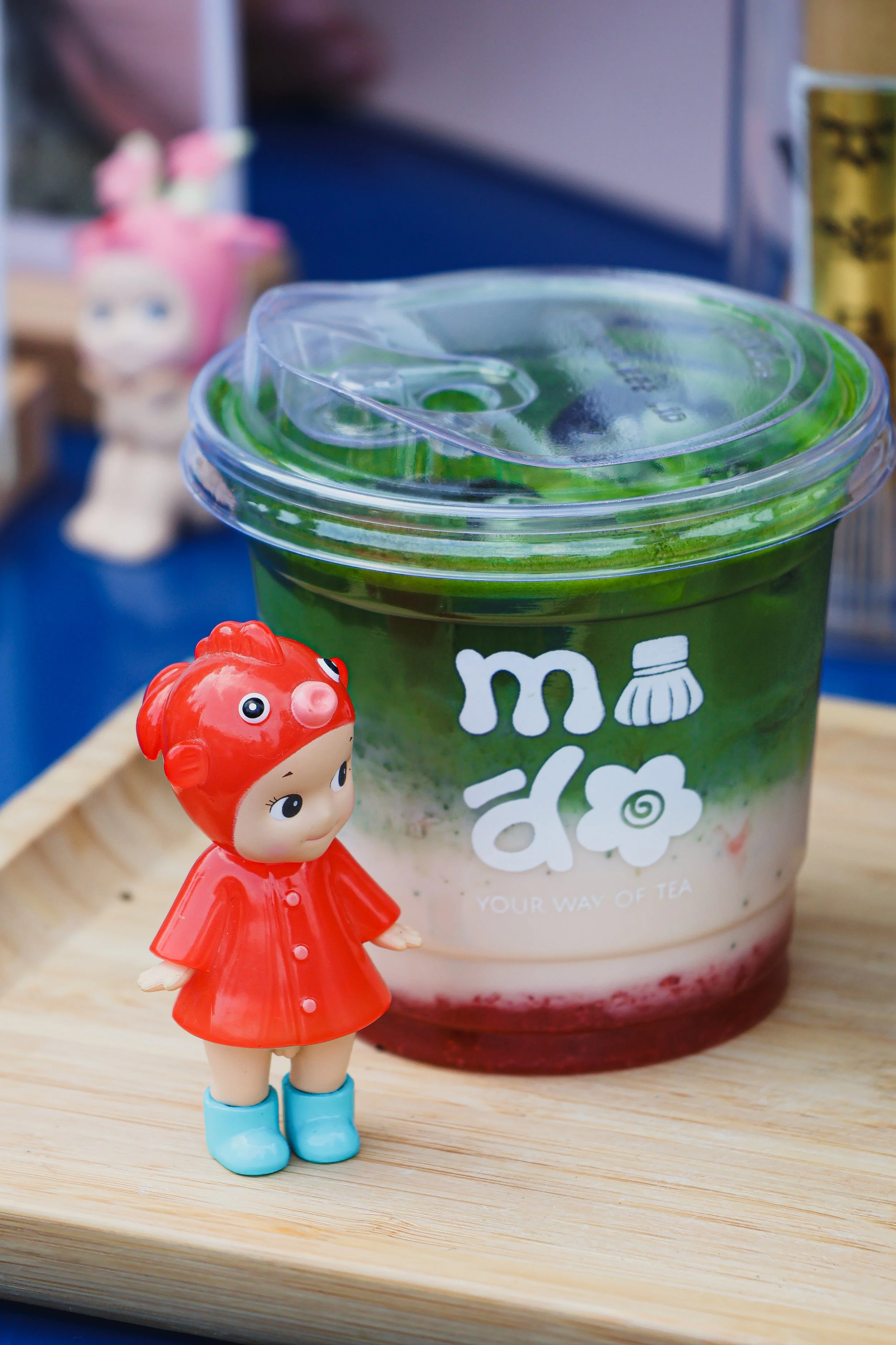 A small toy girl in a red dress and blue boots standing on a wooden surface, next to a plastic cup with a green and white label, and a pink toy character in the background.