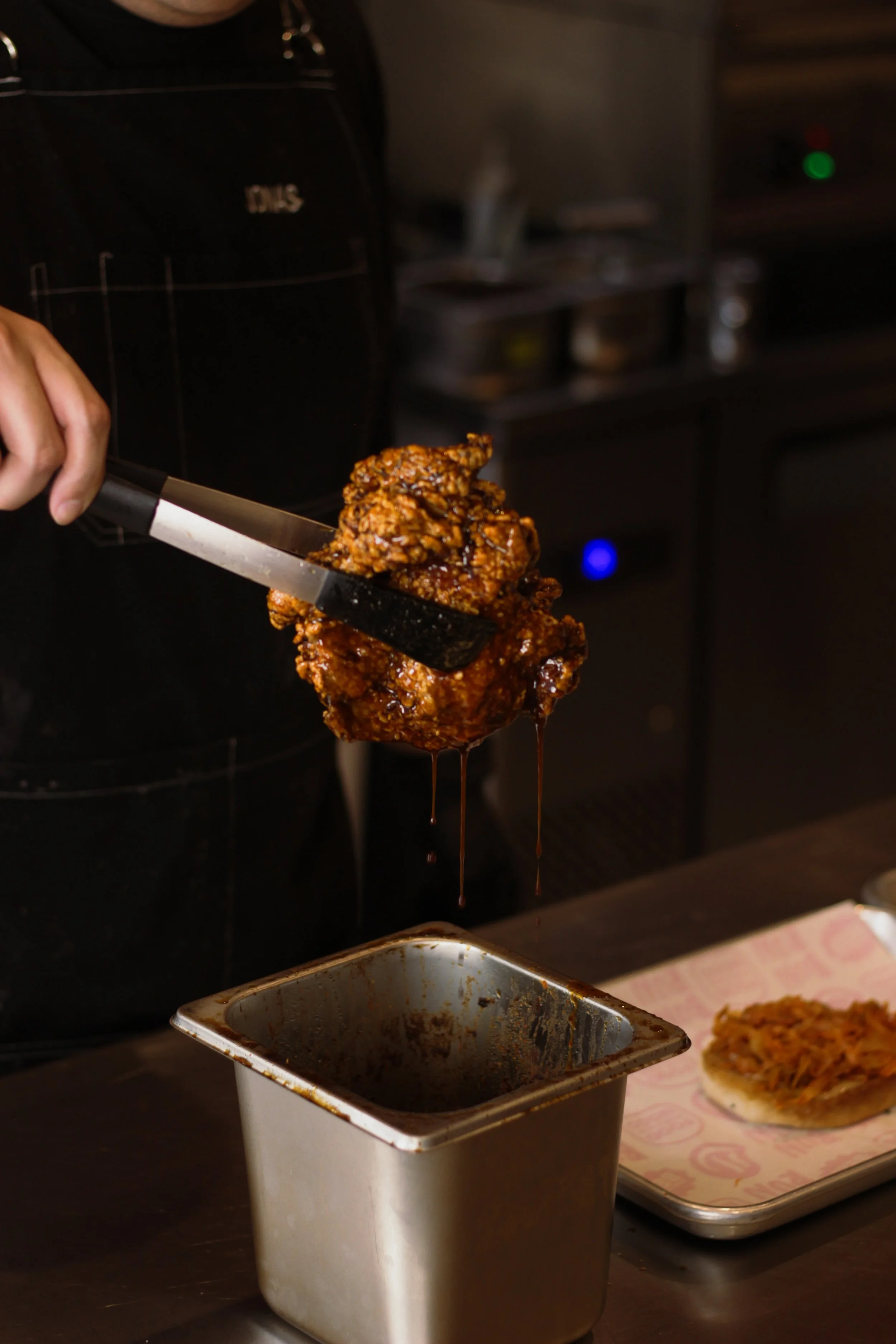 A person using tongs to place a portion of glazed fried chicken into a metal container. A tray with a piece of bread topped with pulled pork or shredded meat in the background.