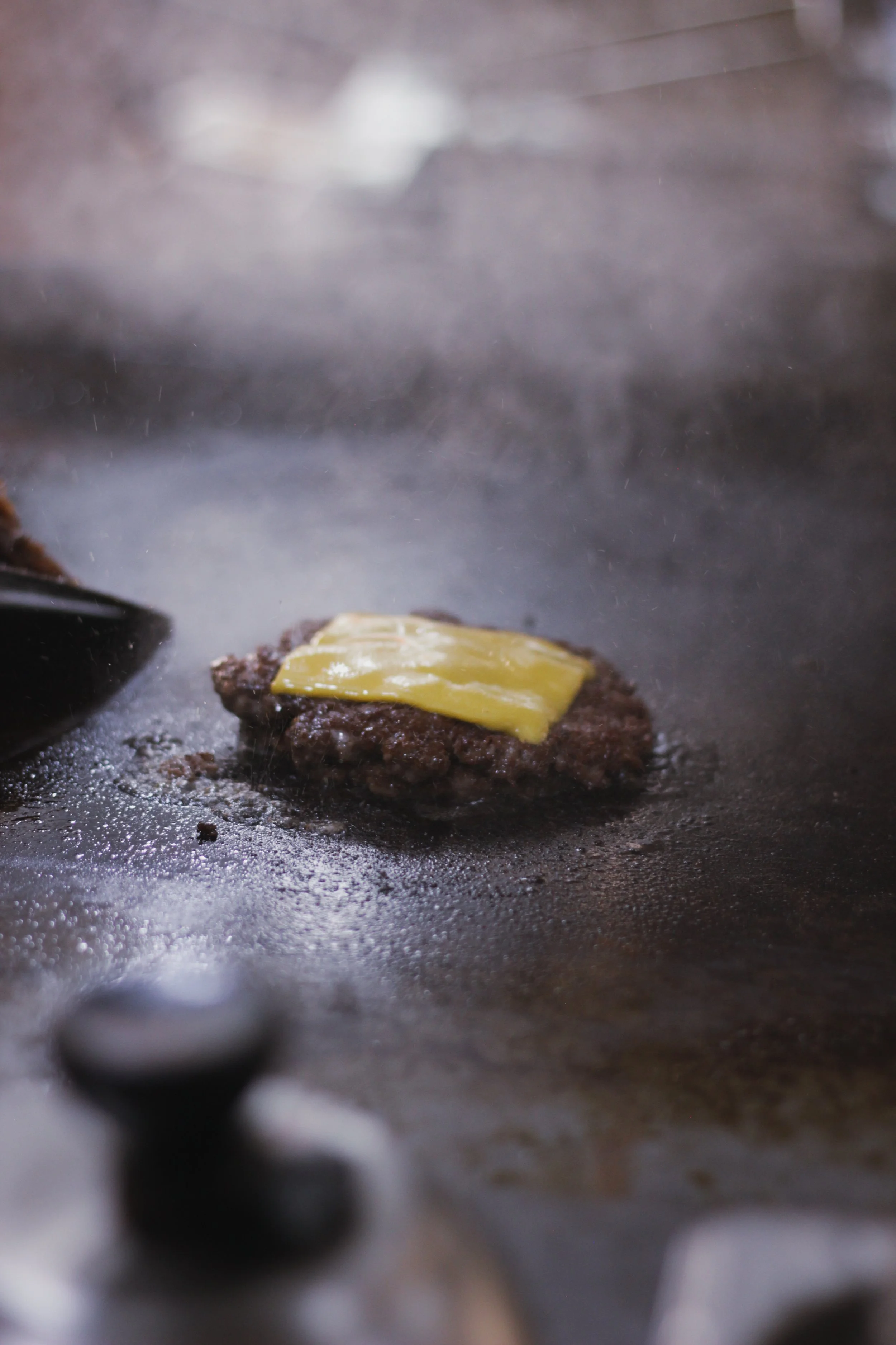 Close-up of a beef patty with melted cheese on top, cooking on a hot griddle.
