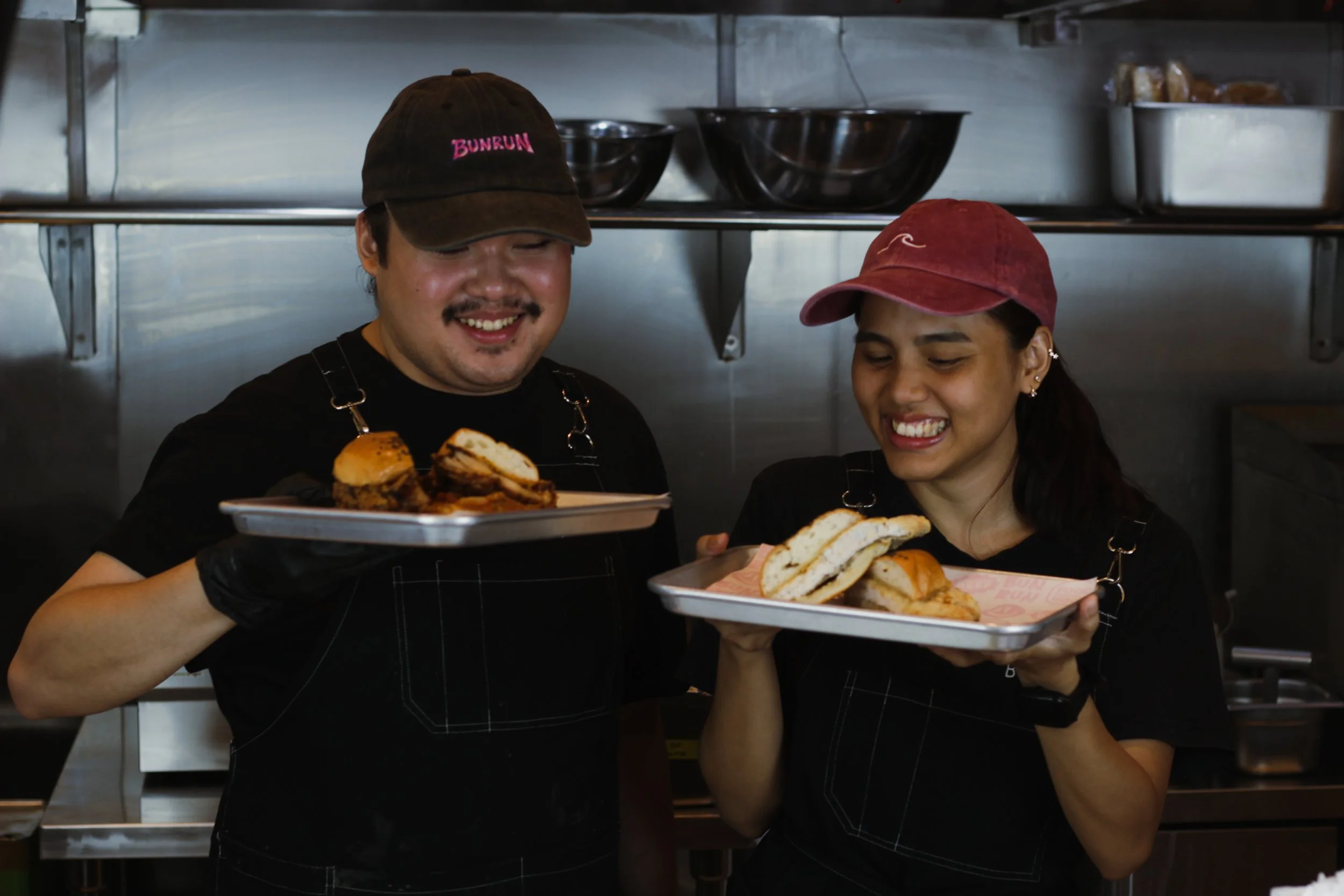 Two smiling food service workers, a man and a woman, holding trays of sandwiches and baked goods in a kitchen.