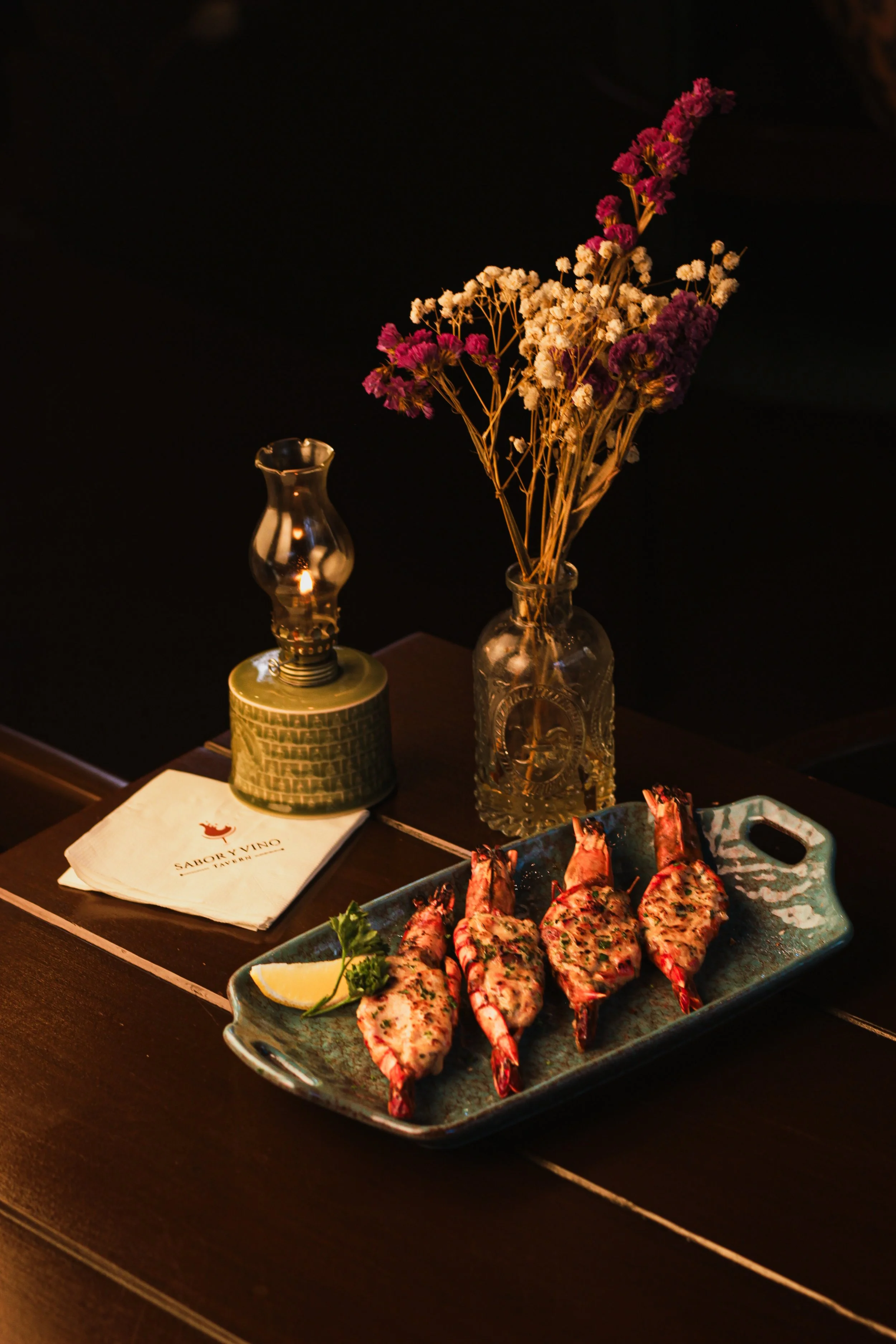 A tray of grilled lobster tails garnished with herbs and lemon slices on a dark table. In the background, there is a vase of pink and white flowers, a small lamp, and a paper napkin with a restaurant logo.
