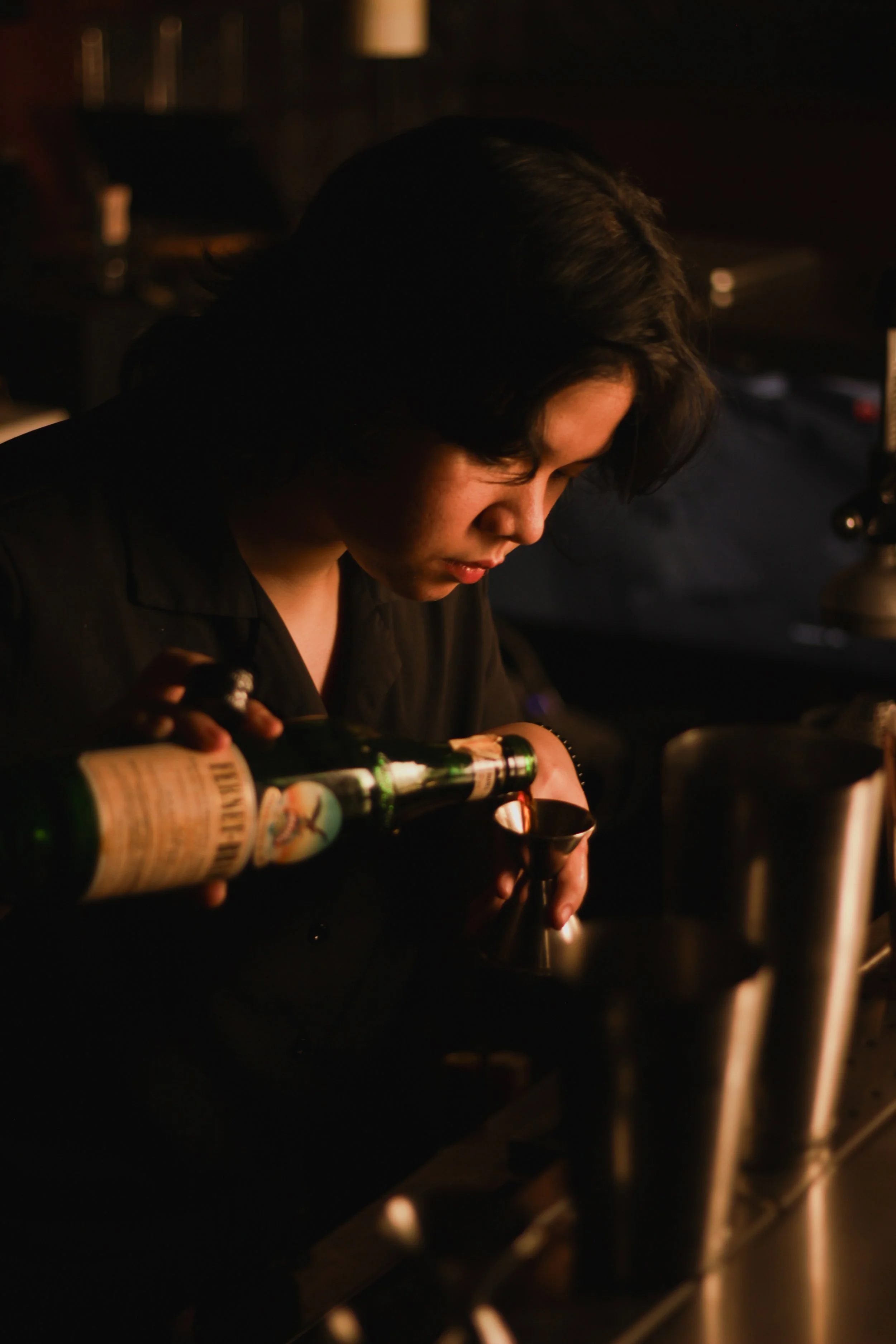 Person with dark hair pouring a drink from a bottle into a metal jigger in a dimly lit setting.