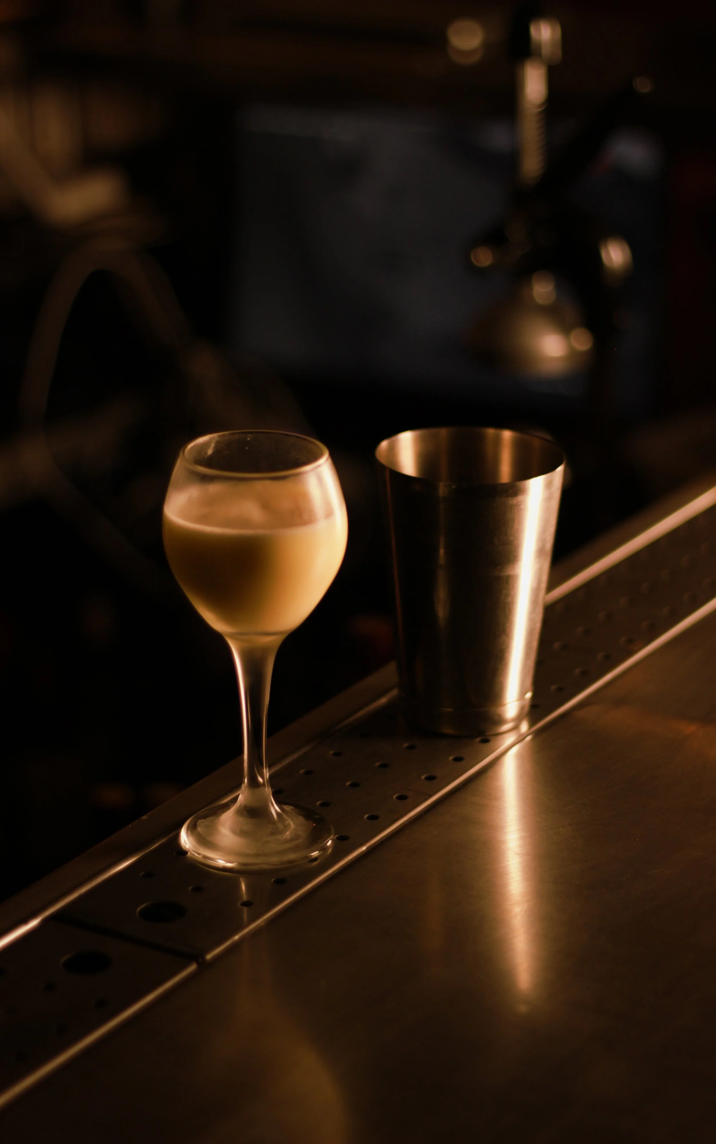 A glass of white wine and a metal shaker on a bar counter with dim lighting.