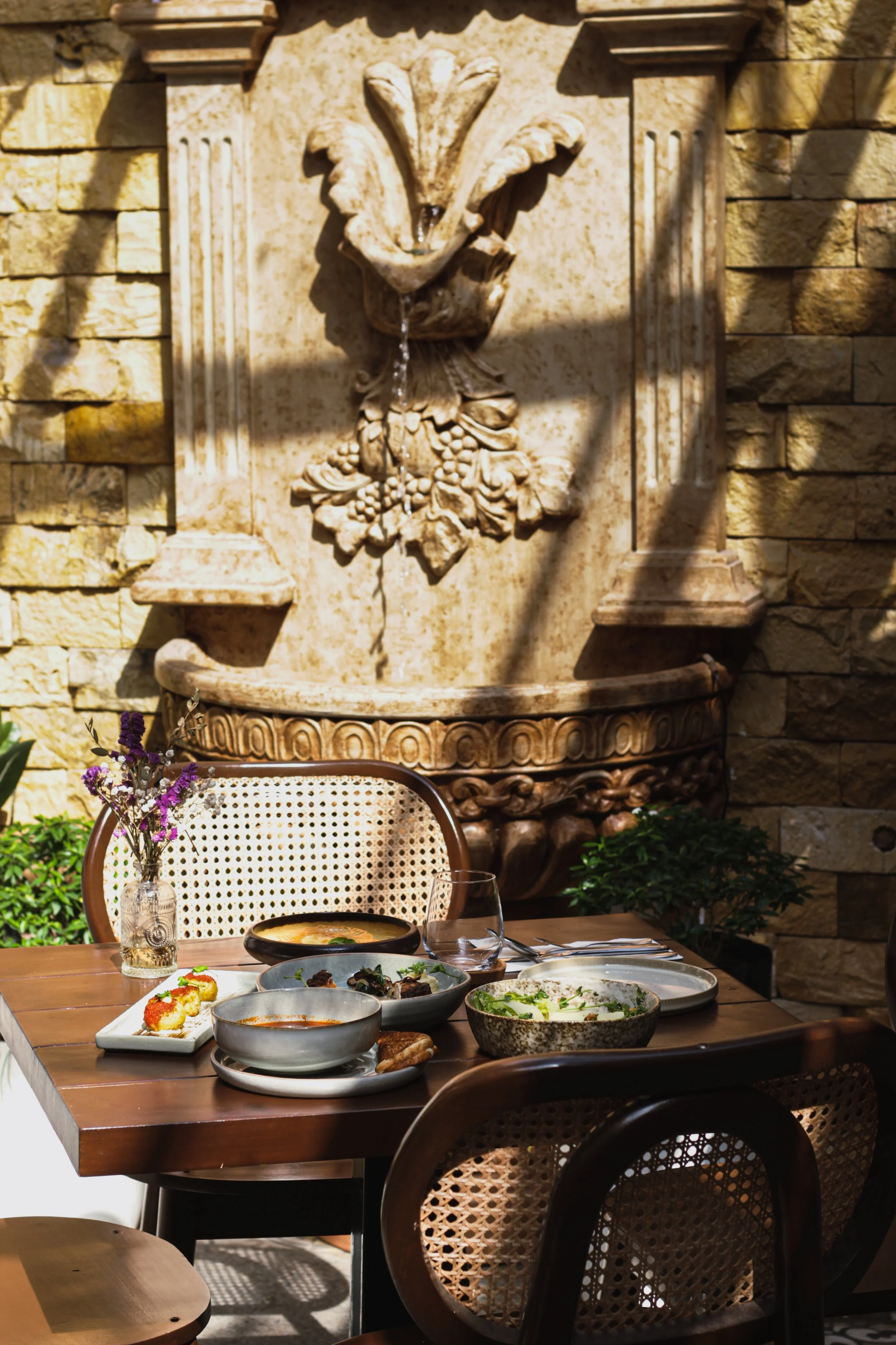 An outdoor dining table set with various dishes, including bowls of soup, salads, and appetizers, on a wooden table. A glass of water and a small vase with purple flowers are also present. A decorative stone fountain with a carved lion's head is mounted on a brick wall in the background. Sunlight casts shadows across the scene.
