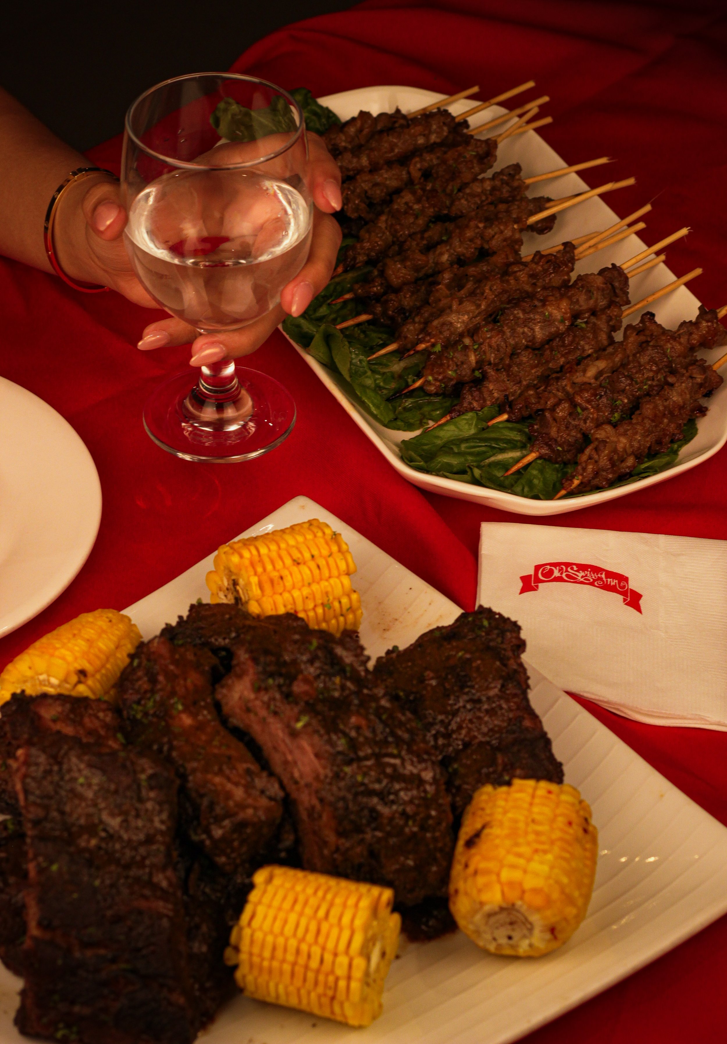 A plate of grilled meat, corn on the cob, a glass of water, and a dish with skewers of grilled meat, with a red cloth background.