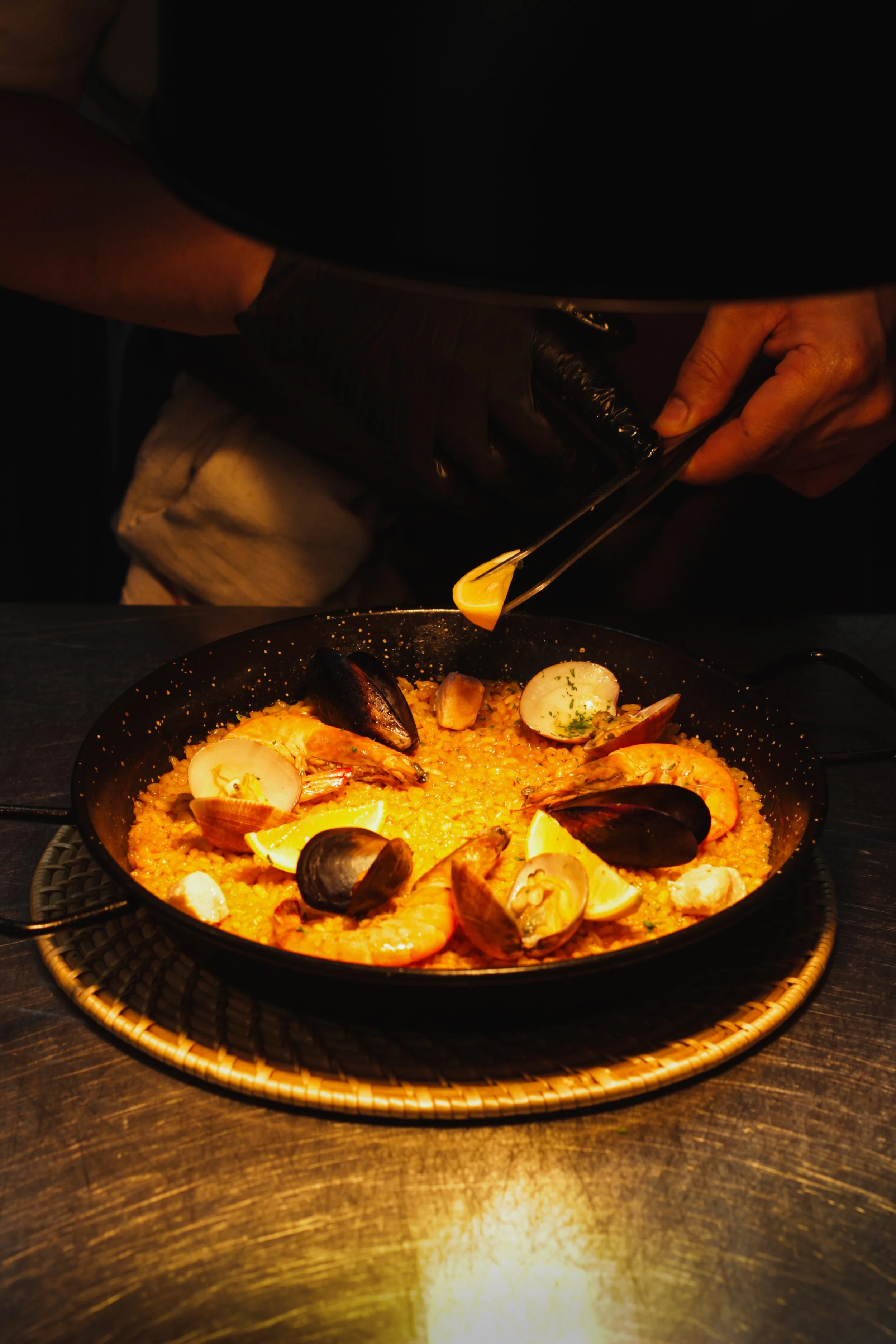 A person wearing black gloves is preparing a seafood paella, which is in a black pan on a round woven placemat, with shrimp, mussels, and lemon wedges. The person is using tongs to add an ingredient to the dish.