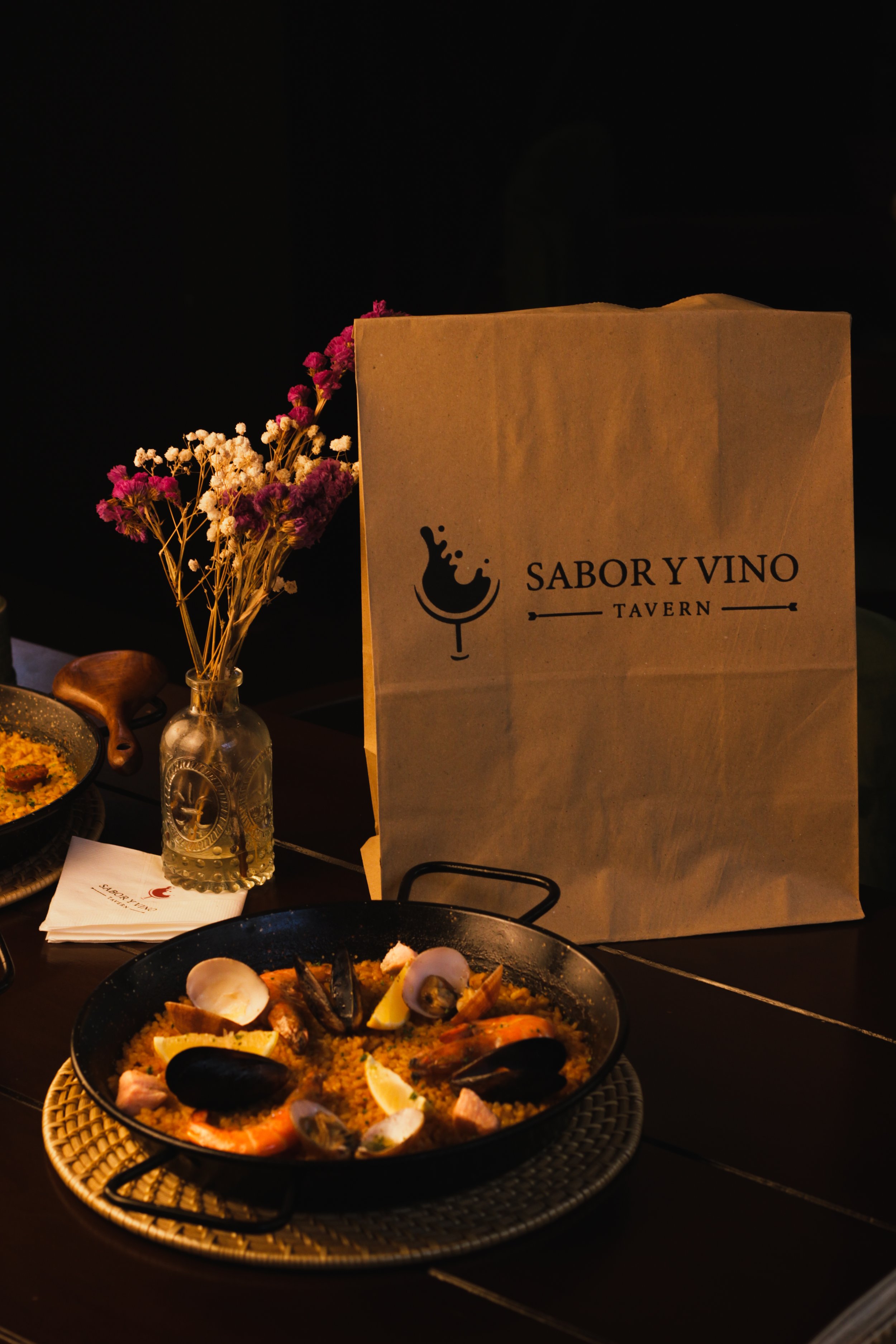 Plate of seafood paella with shrimp, mussels, and lemon slices, with a floral arrangement and a paper bag from Sabor y Vino Tavern on a dark table.