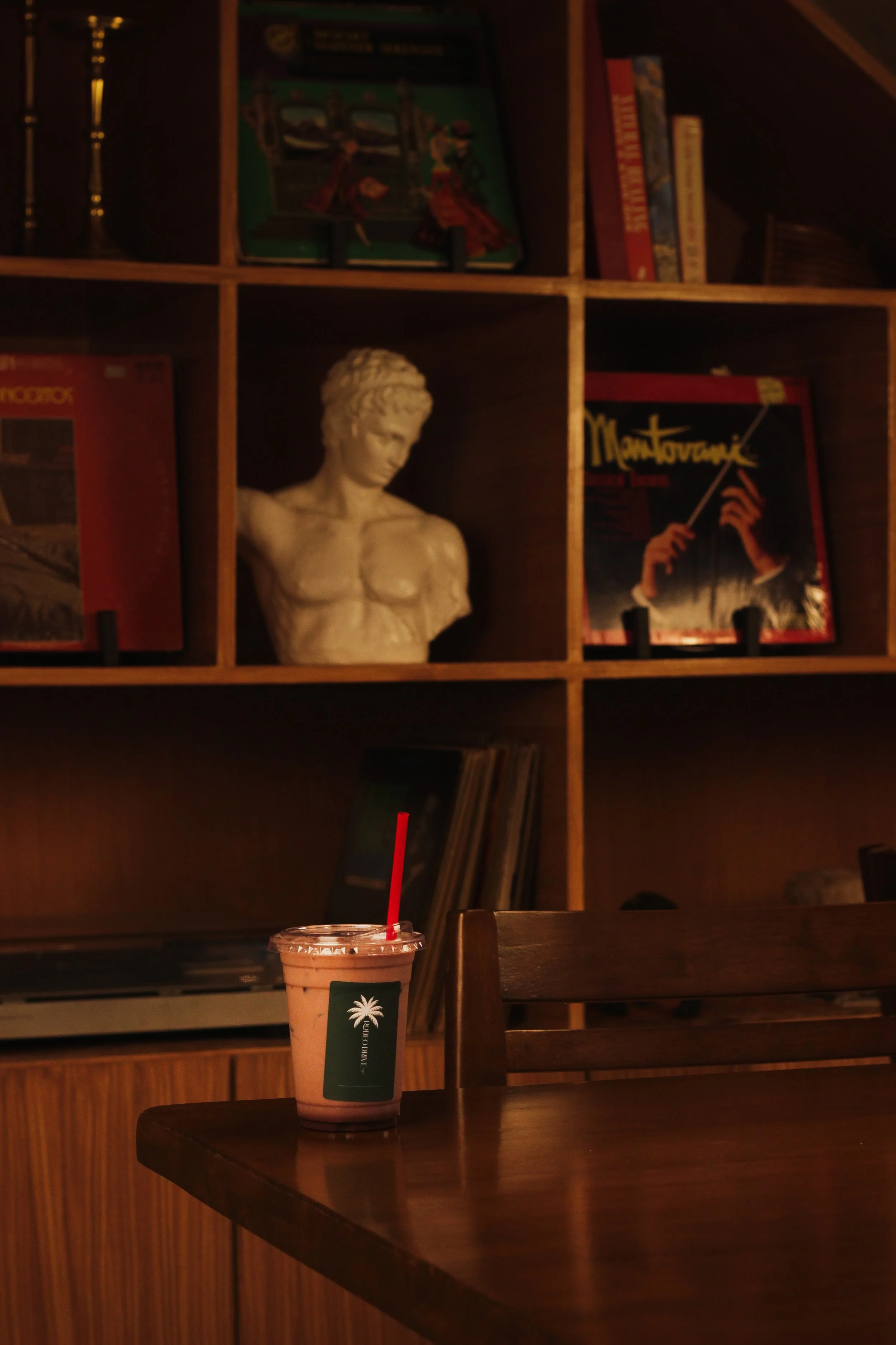 A wooden table with a plastic cup containing a pink beverage and a red straw, in front of a wooden bookshelf with books and decorative items, including a white bust sculpture.