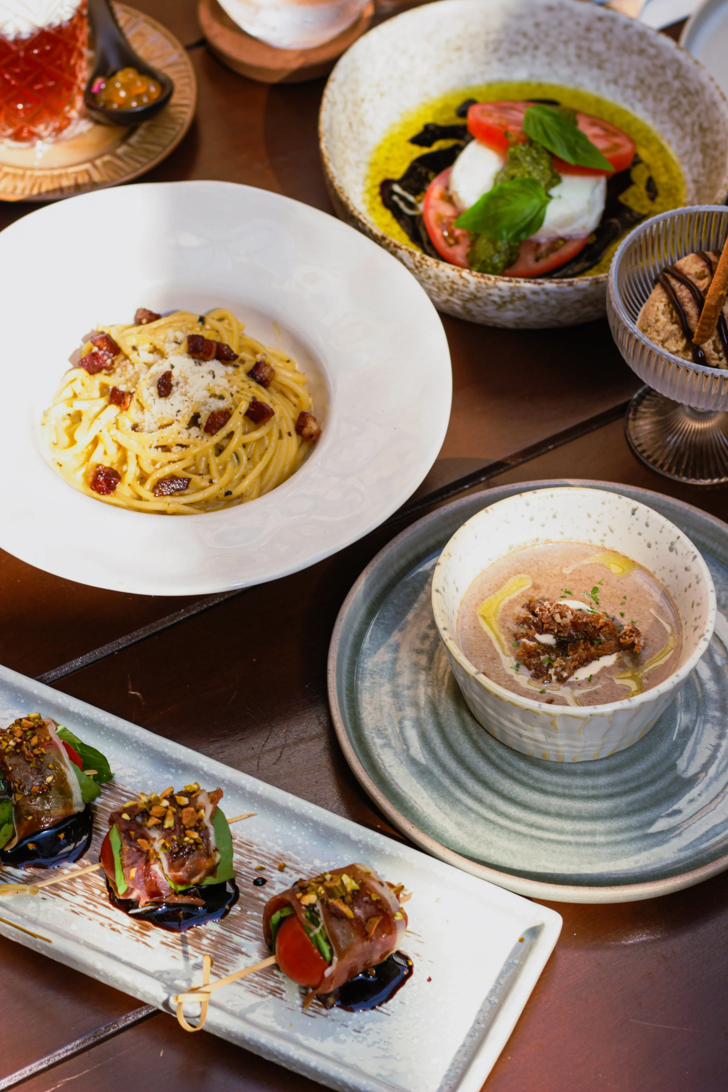 A variety of gourmet dishes including a plate of spaghetti with bacon, a bowl of Caprese salad with tomatoes, mozzarella, basil, and olive oil, a bowl of creamy soup, and a tray of skewered appetizers garnished with chopped nuts and drizzled sauces.