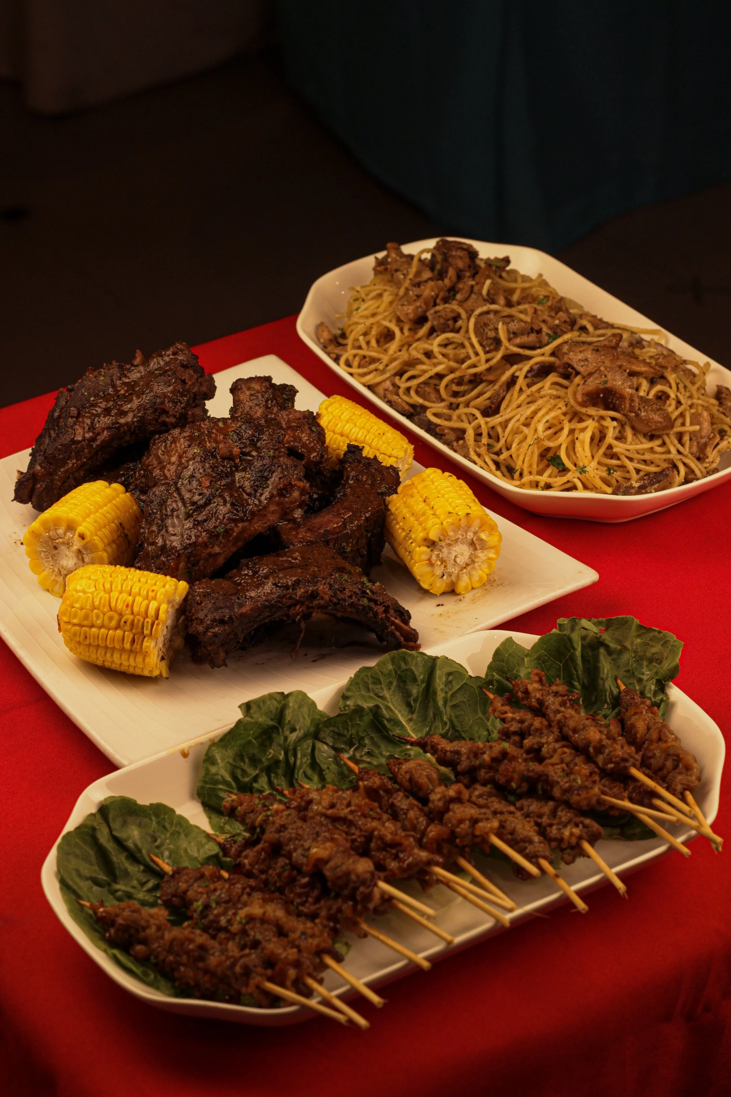 A variety of dishes on a red tablecloth, including grilled meats with corn on the cob, spaghetti with beef, and skewered beef served with green leafy garnish.