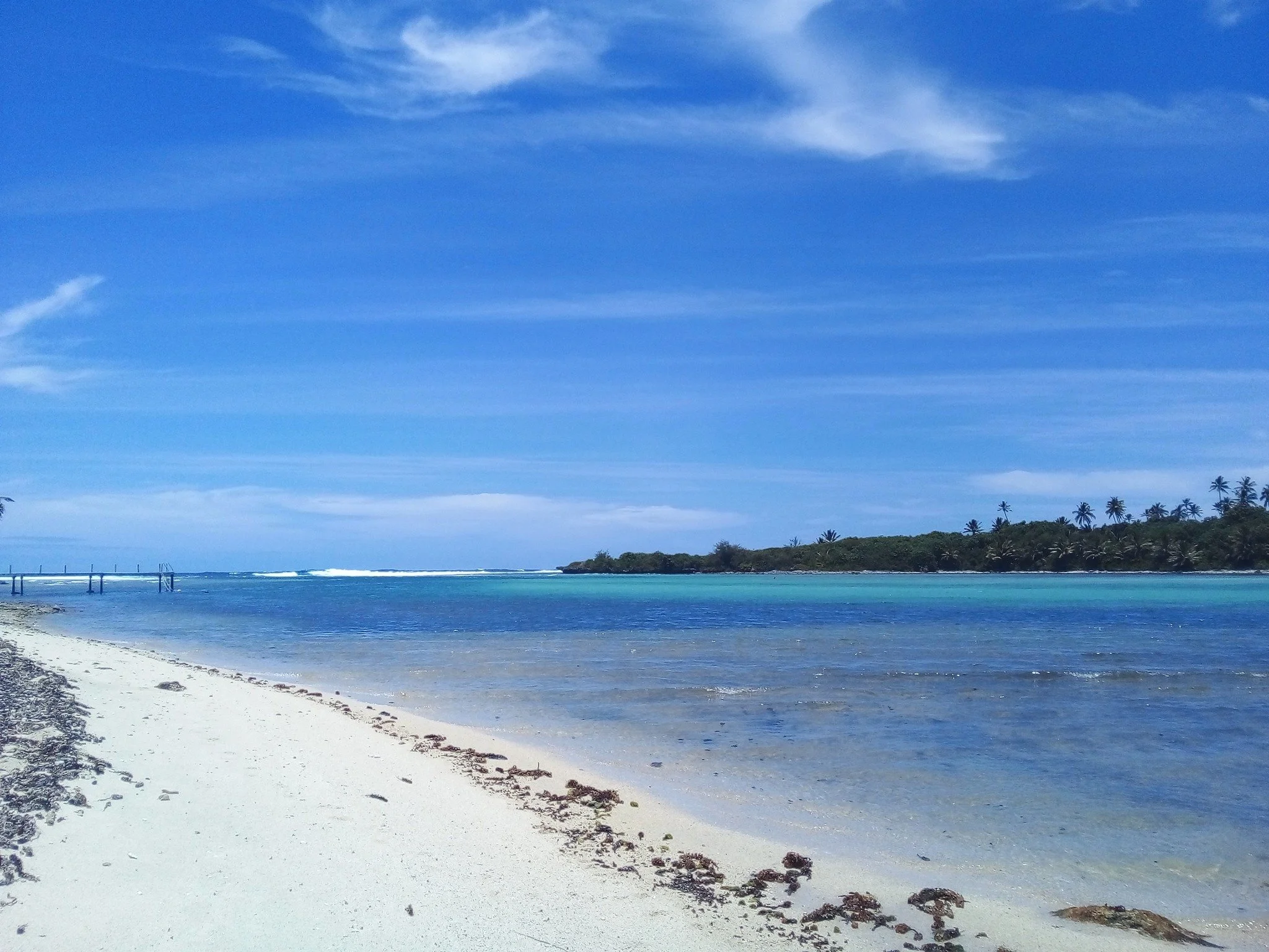 A beach at Avana Passage, Rarotonga, Cook Islands, with white sand, clear blue water, and a line of palm trees in the background under a mostly cloudy sky.