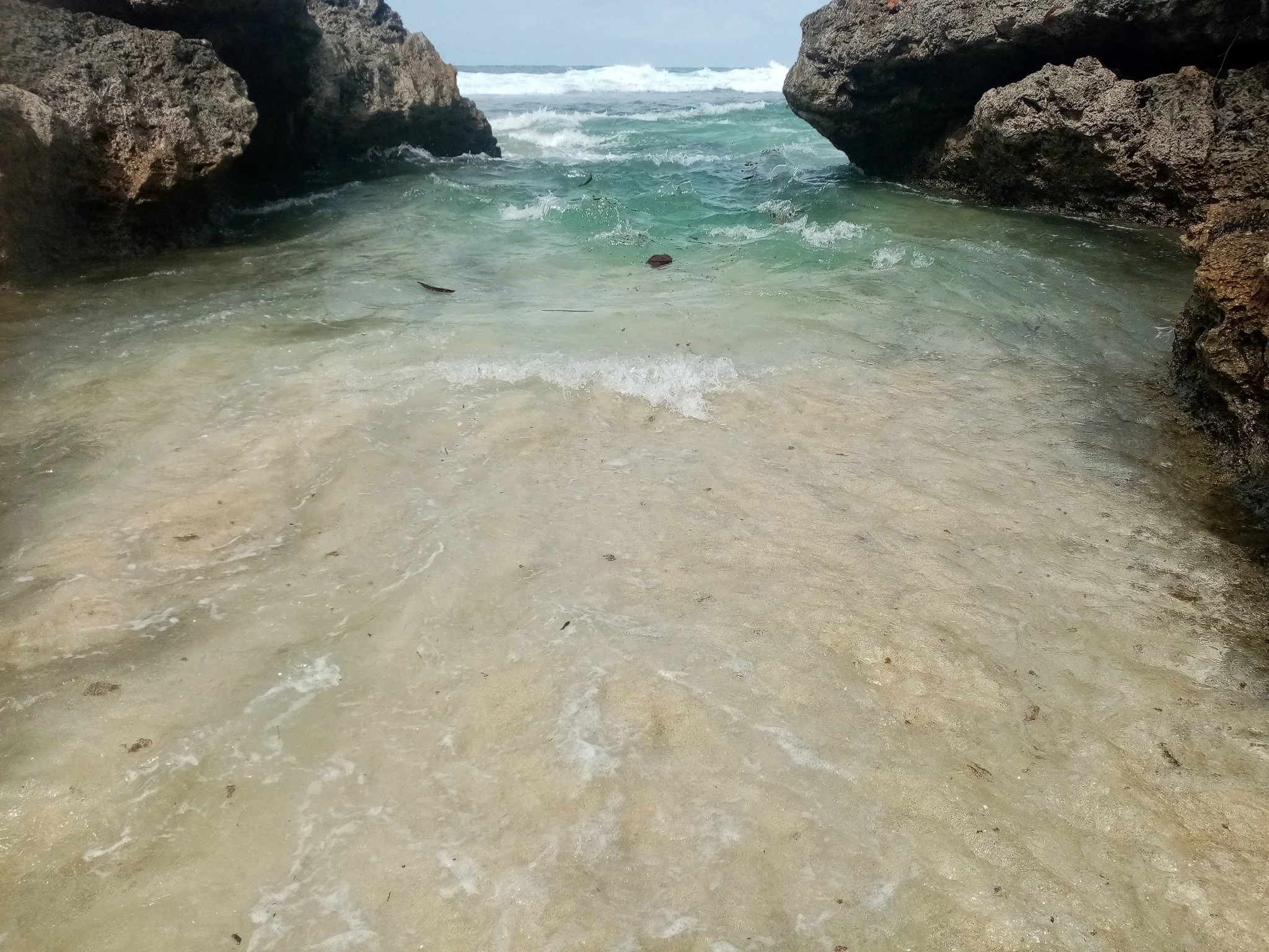 The image shows a view of the ocean through a natural rock formation, with clear turquoise water and white waves crashing in the background, and sandy beach in the foreground. Location Rarotonga Cook Islands