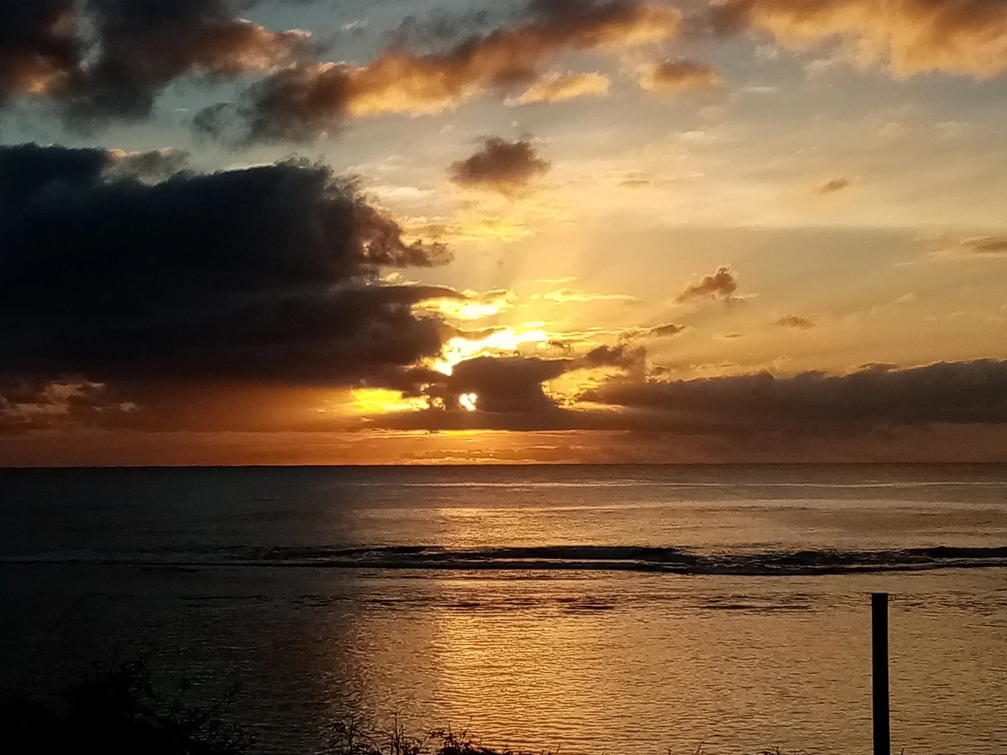 A sunset over the ocean at Black Rock Rarotonga, Cook Islands, with clouds in the sky reflecting the golden and dark hues of the setting sun.