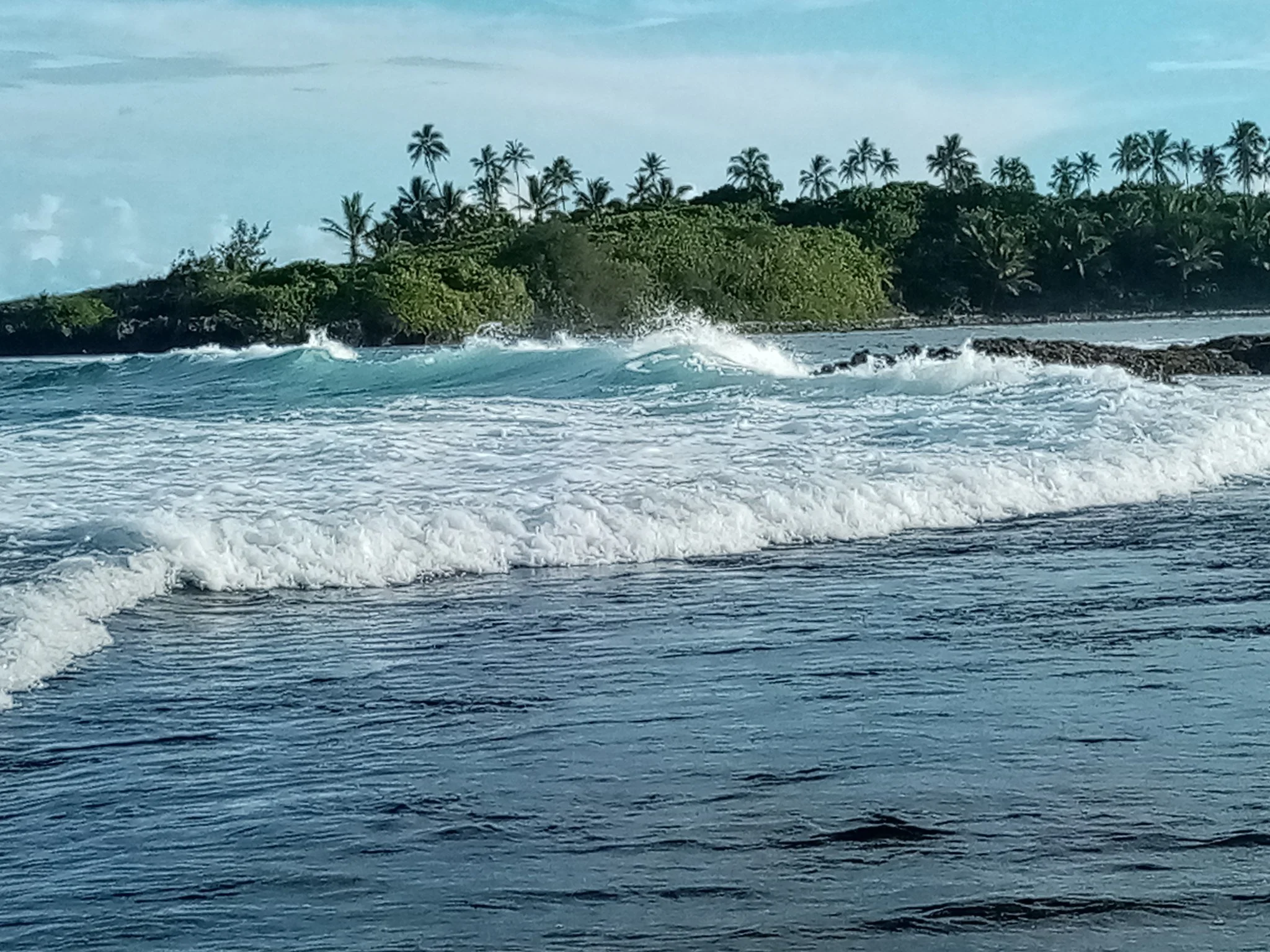 Avana Passage, Rarotonga, Cook Islands, where vaka, canoes left for Aotearoa, New Zealand, Ocean waves crashing against a rocky shoreline with lush green tropical trees and palm trees in the background under a partly cloudy sky.