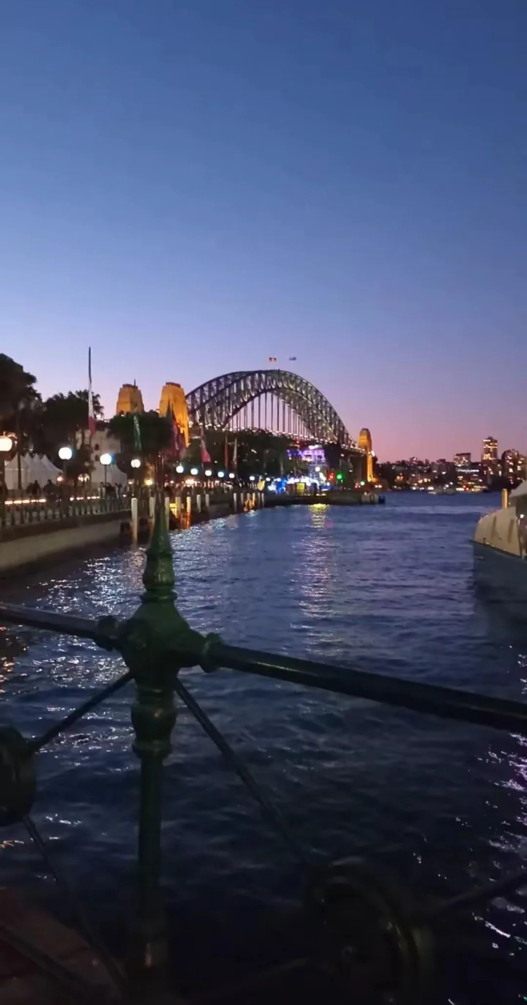 Evening view of the Sydney Harbour Bridge in Australia, lit up with colorful lights, over a waterway with reflections, and a promenade with street lamps and people walking.