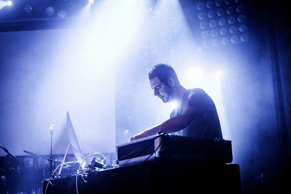 A DJ performing on stage with equipment, backlit by bright blue and white lights.