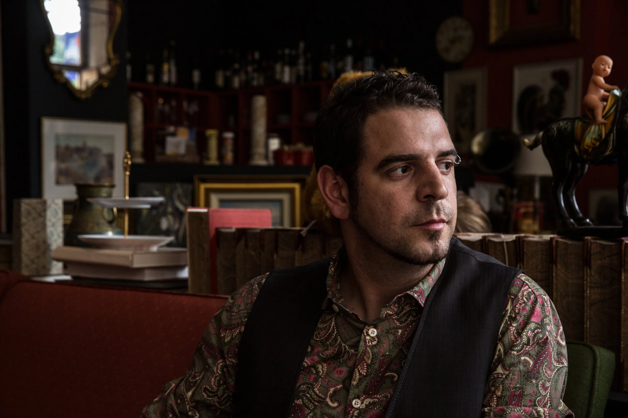 A man with dark hair, a patterned shirt, and a dark vest sitting in a dimly lit room decorated with artwork, a mirror, and various decorative objects.
