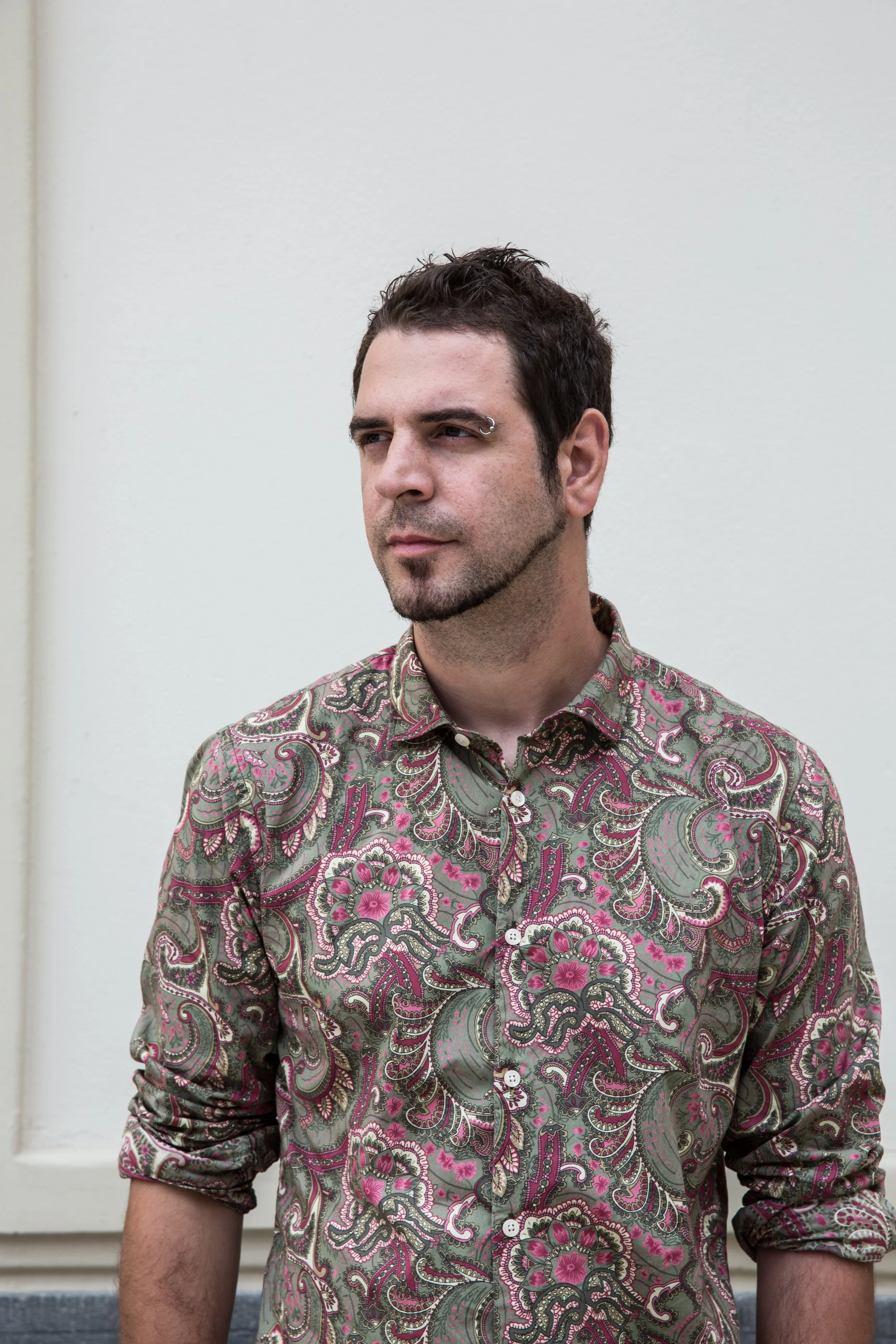 A man with dark hair, facial hair, and a metal eyebrow ring, wearing a colorful paisley patterned shirt with rolled-up sleeves, standing against a plain white wall.