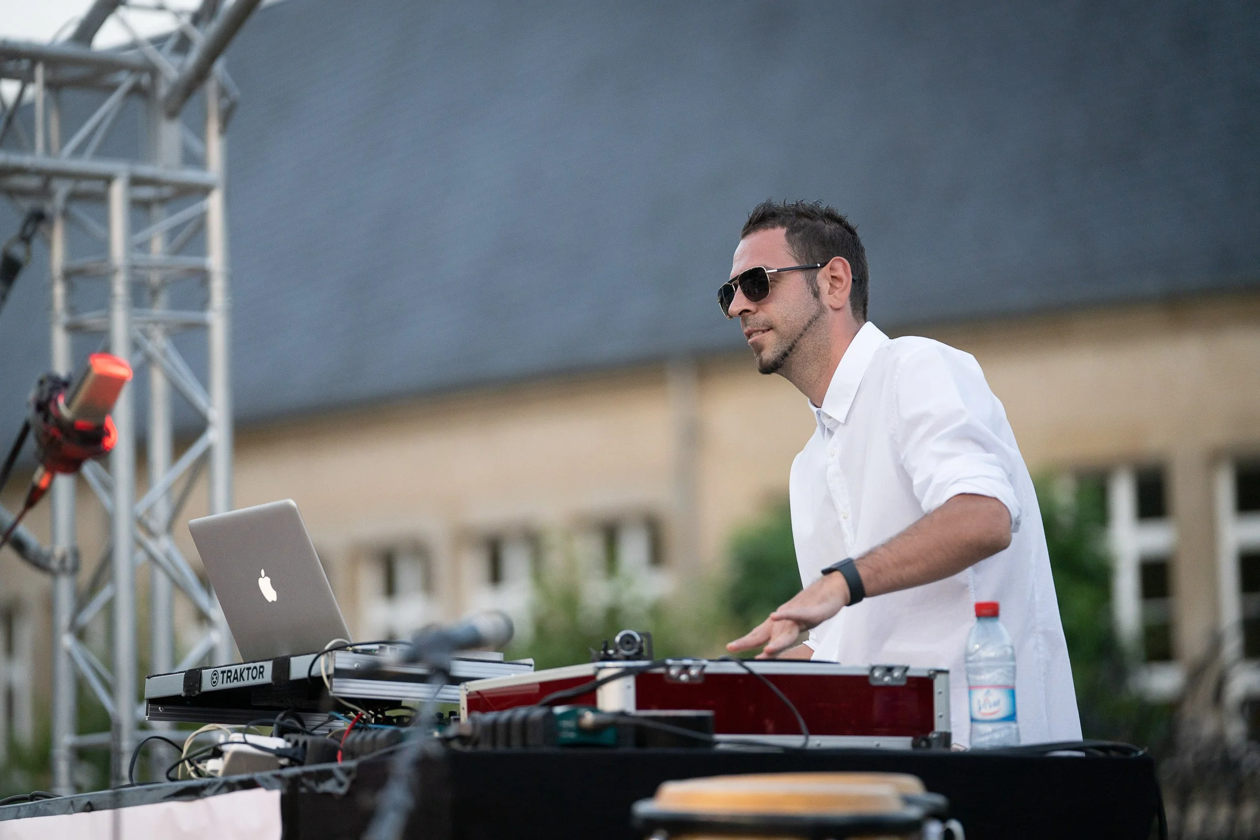 A man in sunglasses and a white shirt DJing outdoors with music equipment and a laptop.
