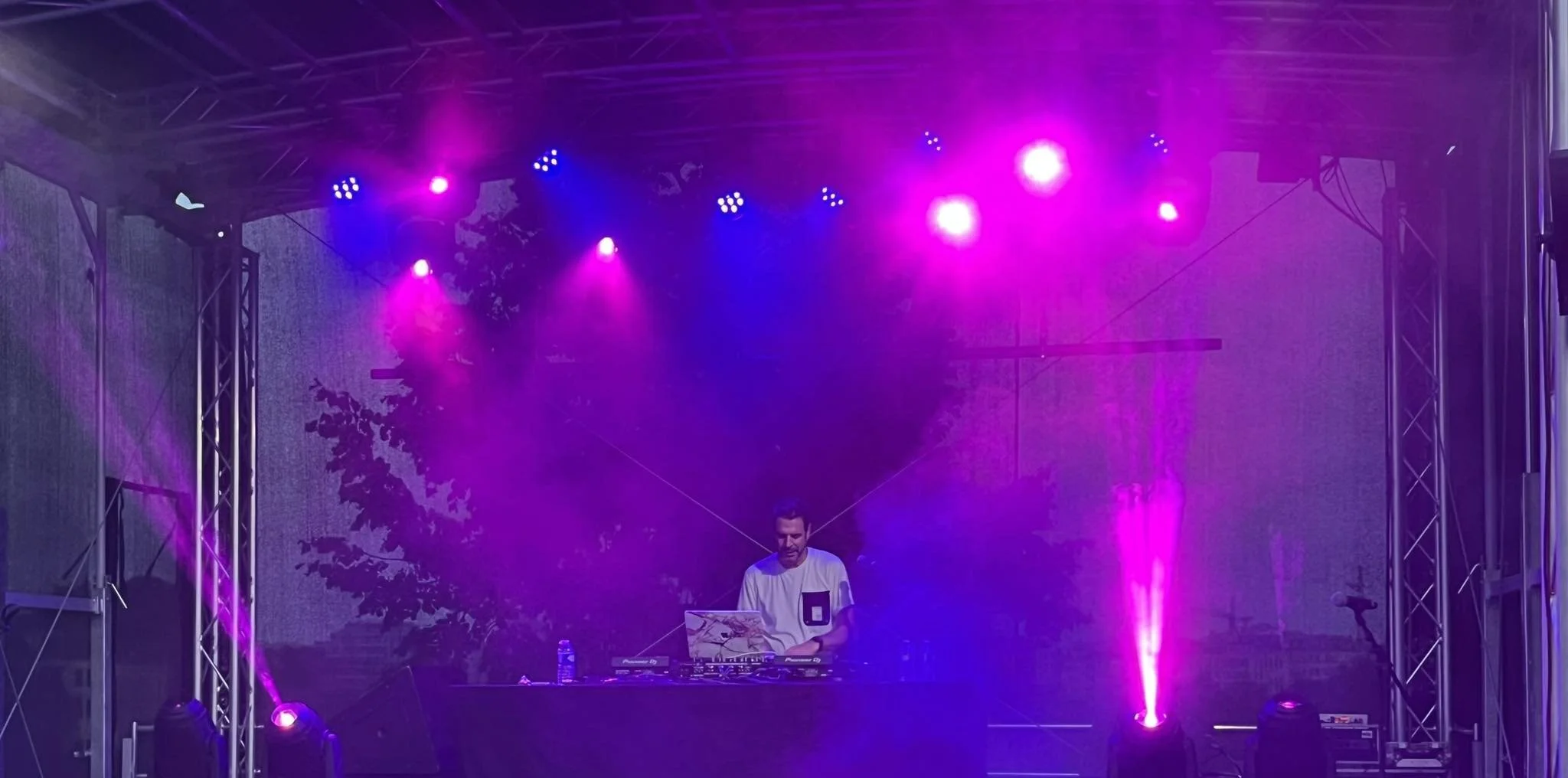 A DJ performing on a stage illuminated with purple and pink lights, with a backdrop of trees and outdoor surroundings.