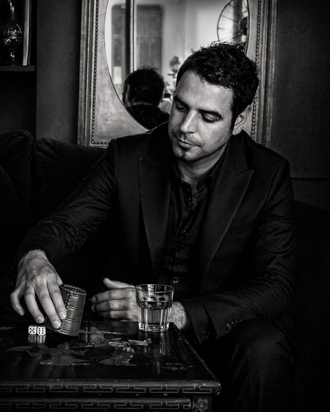A man in a black suit sitting at a table with a glass of water, playing dominoes, in a room with a large mirror behind him.