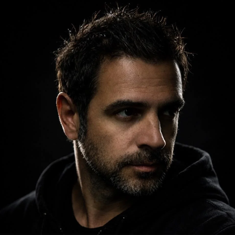 Close-up portrait of a man with dark hair and beard, looking to the side against a black background.