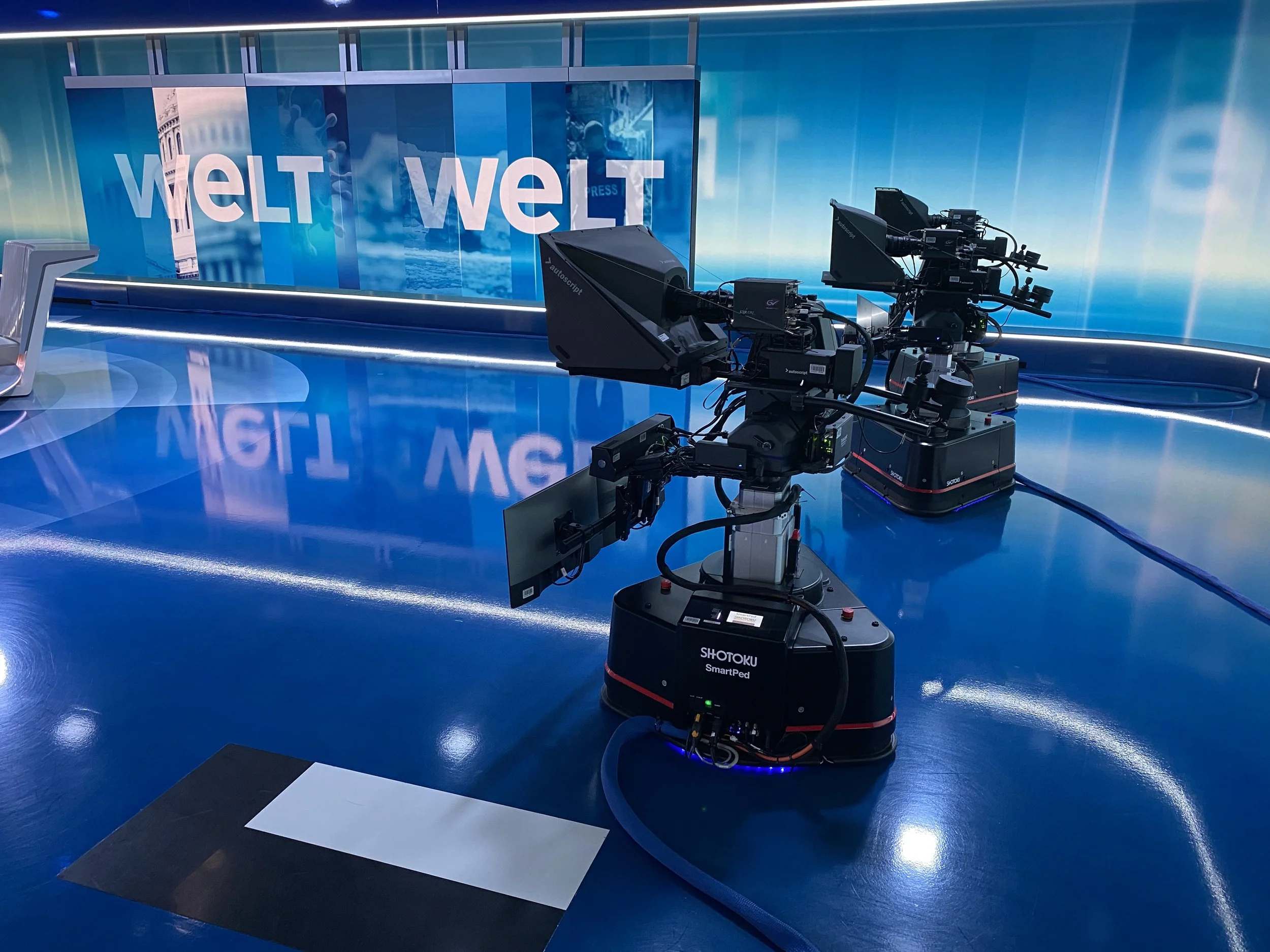A television studio with a blue and black stage floor, featuring professional camera equipment on robotic dollies. The background displays a large screen with the word 'WELT' in white letters.