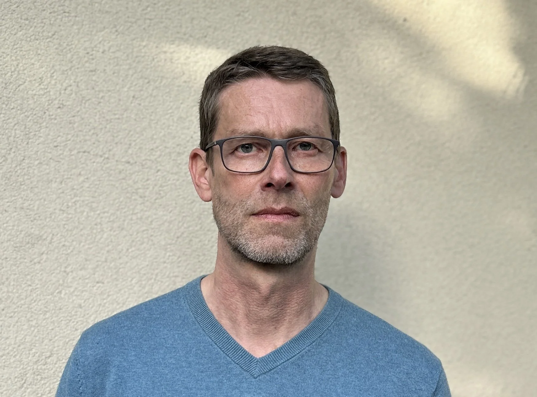 A middle-aged man with short brown hair, glasses, and light facial hair, wearing a blue V-neck sweater, standing against a beige wall.