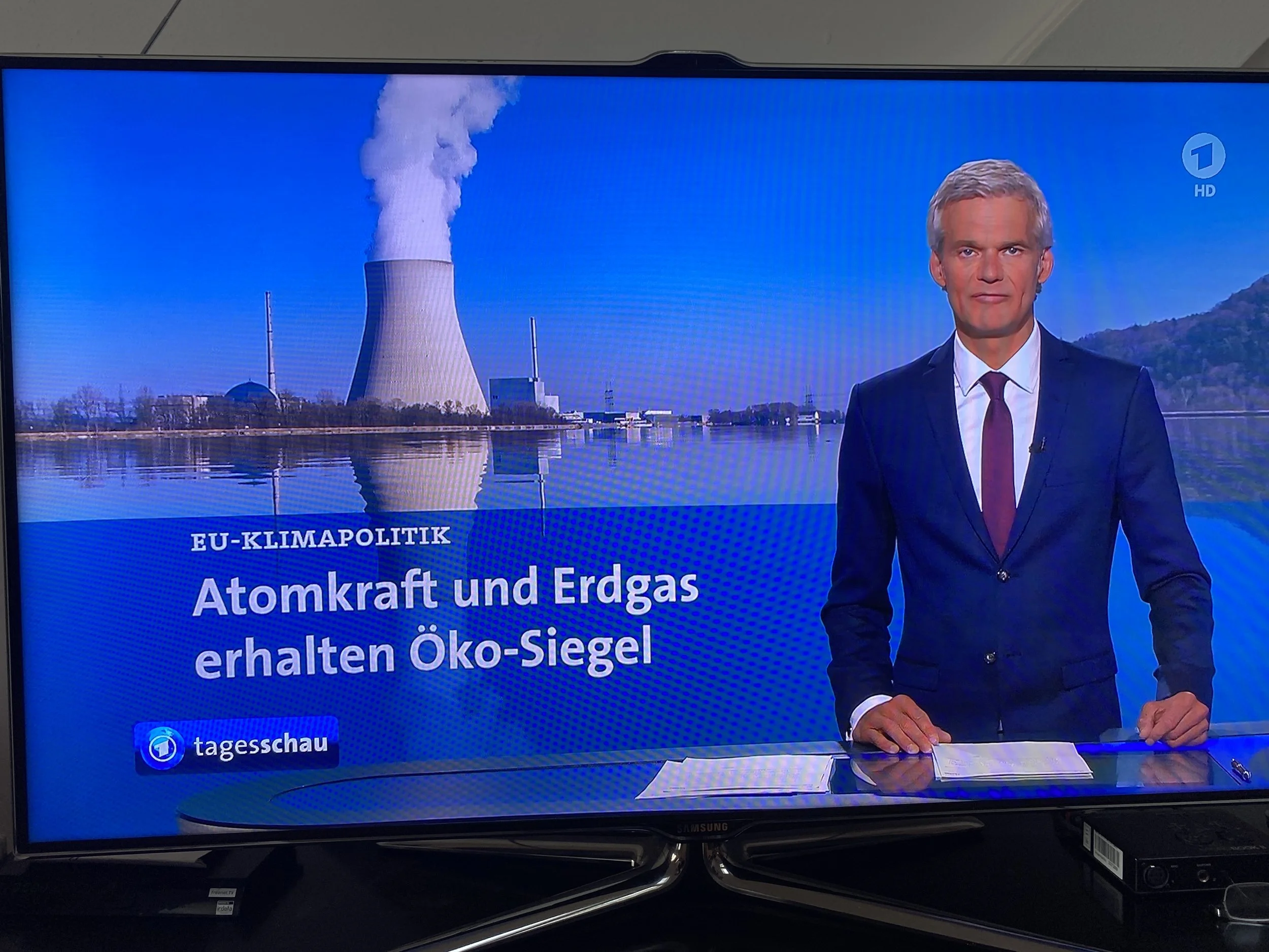A television news broadcast shows a man in a suit and tie standing at a news desk with a scenic backdrop of a nuclear power plant emitting steam or smoke, and a blue sky. The screen has text in German about nuclear power and fossil fuels.