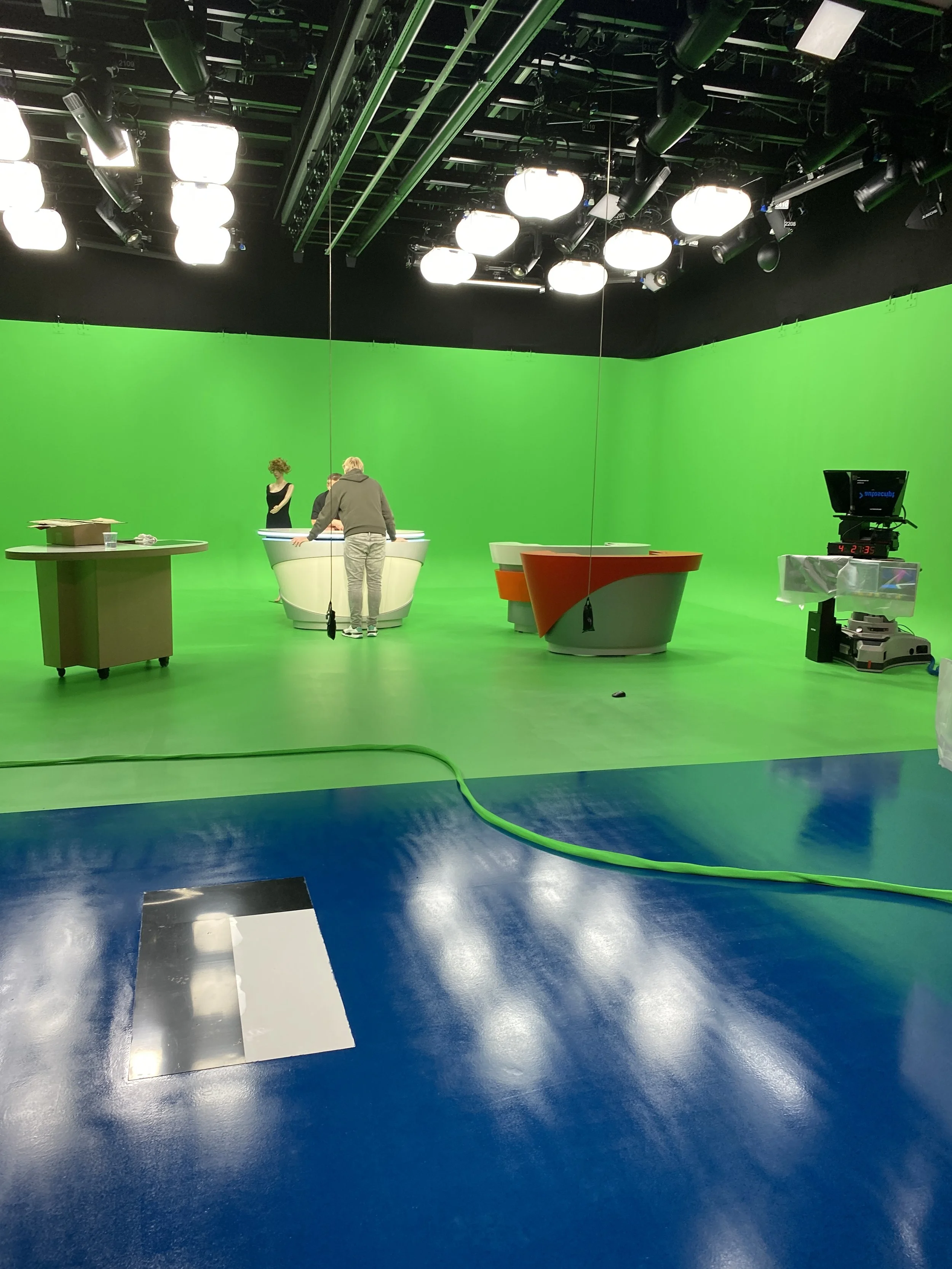 A television studio with a green screen background and broadcast equipment. Three people are gathered around a table in the studio.
