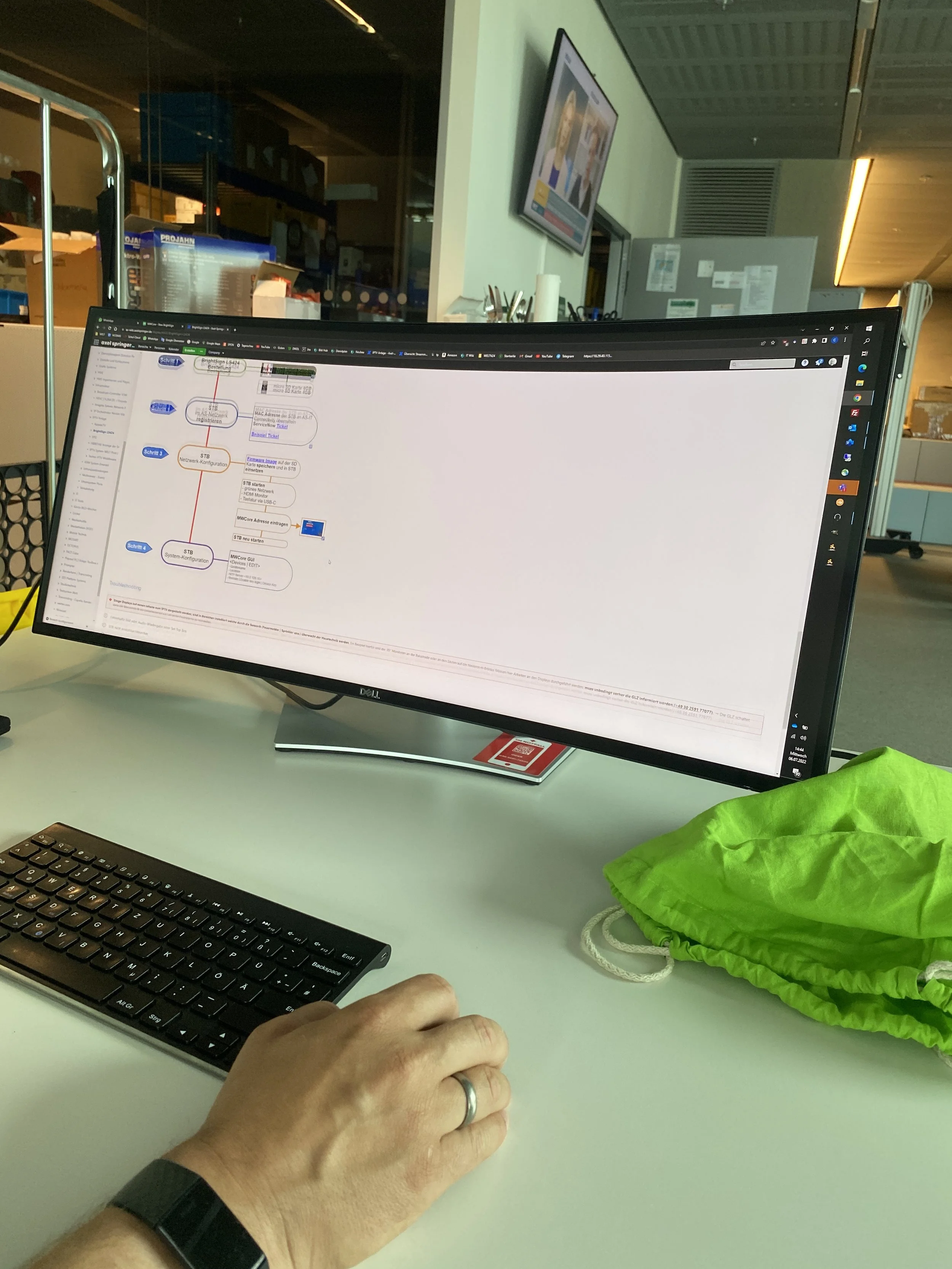 A computer monitor displays a flowchart or diagram in a workspace or office environment. A person's hand is on a keyboard, and there is a green drawstring bag on the desk. In the background, a monitor on the wall shows a video call or presentation.