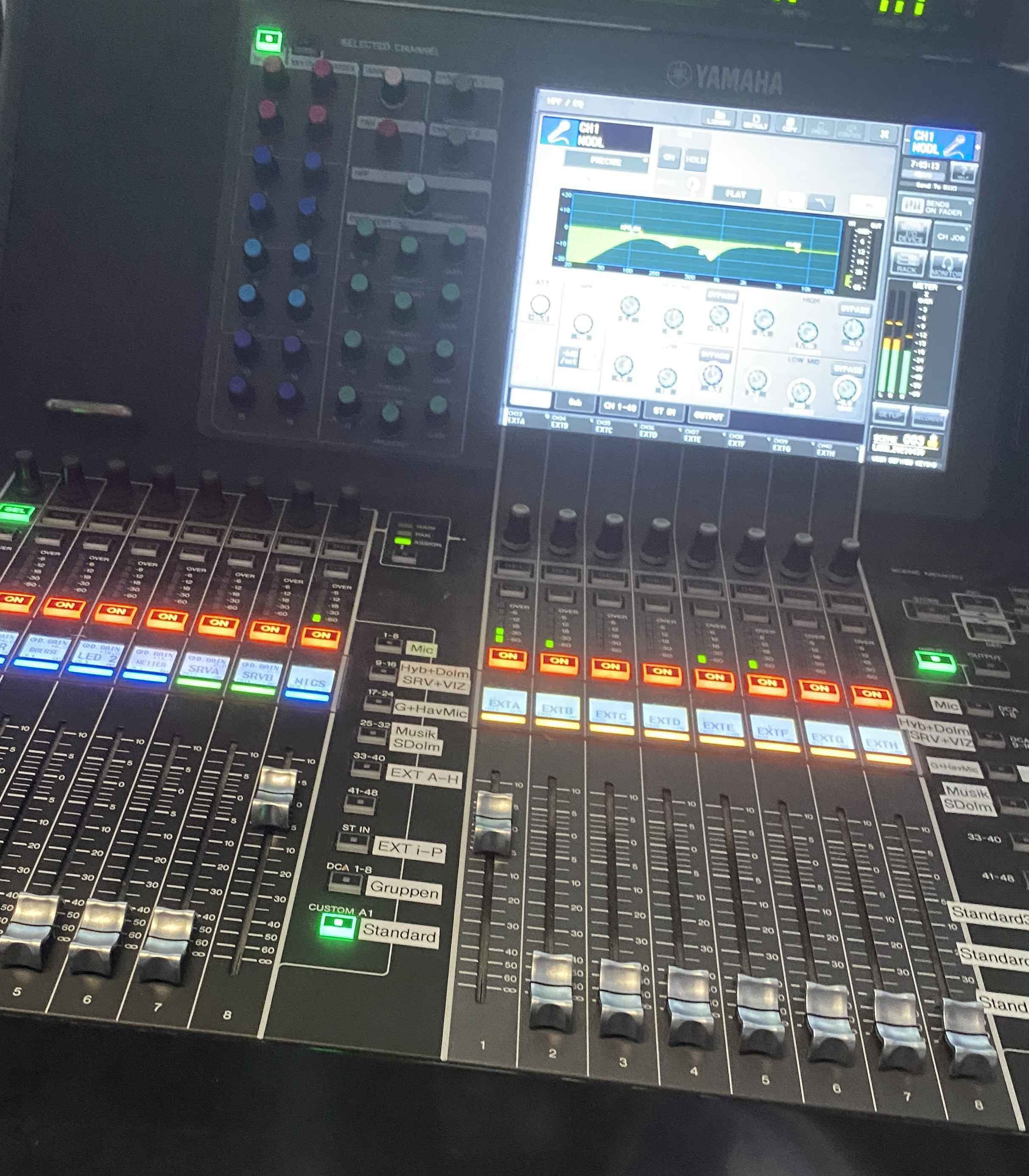 Close-up view of a Yamaha audio mixing console with various knobs, sliders, and a digital screen displaying an equalizer graph and controls.