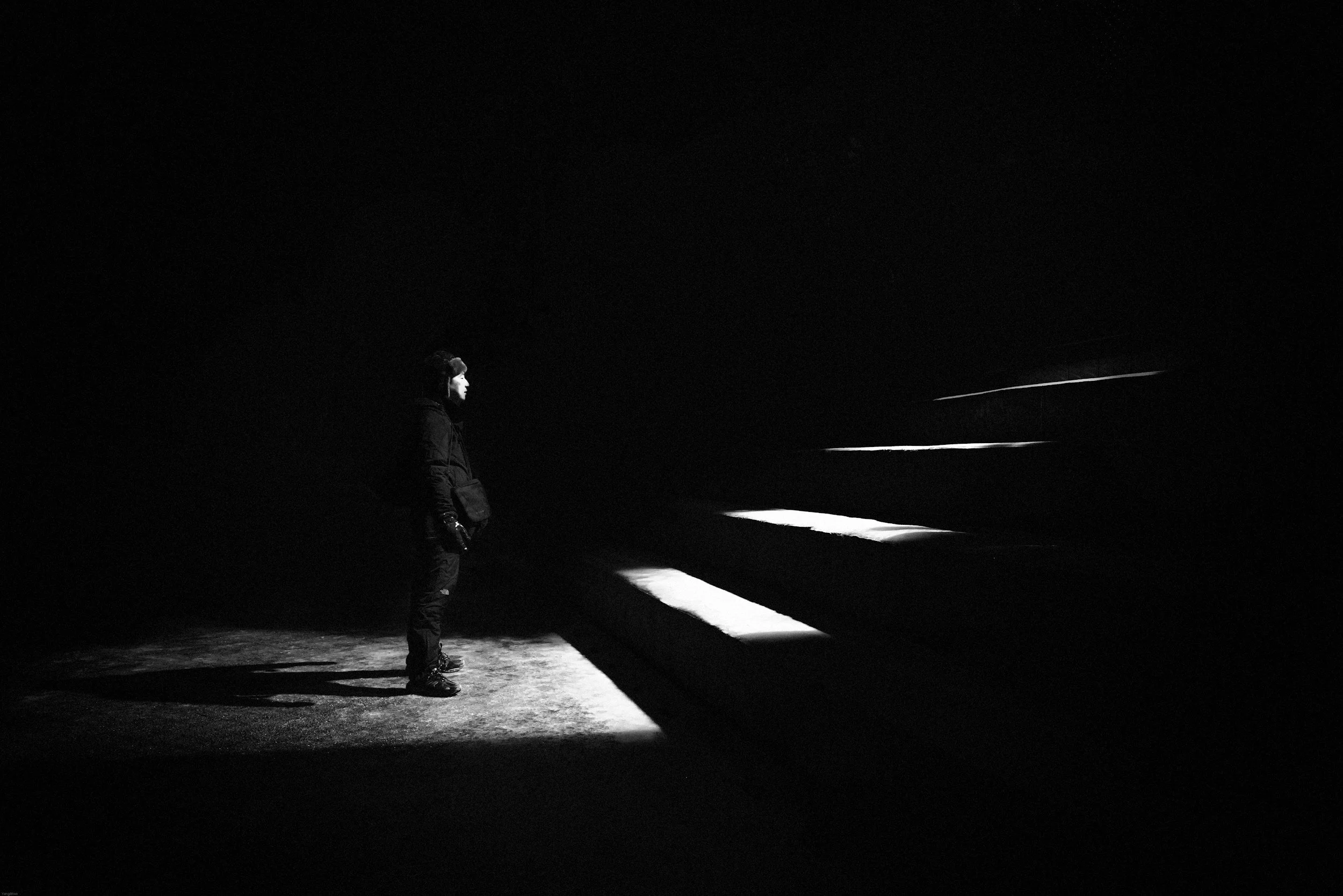 A person standing outdoors at night, illuminated by a nearby light source, looking at a dark landscape with faint light streaks across the sky.