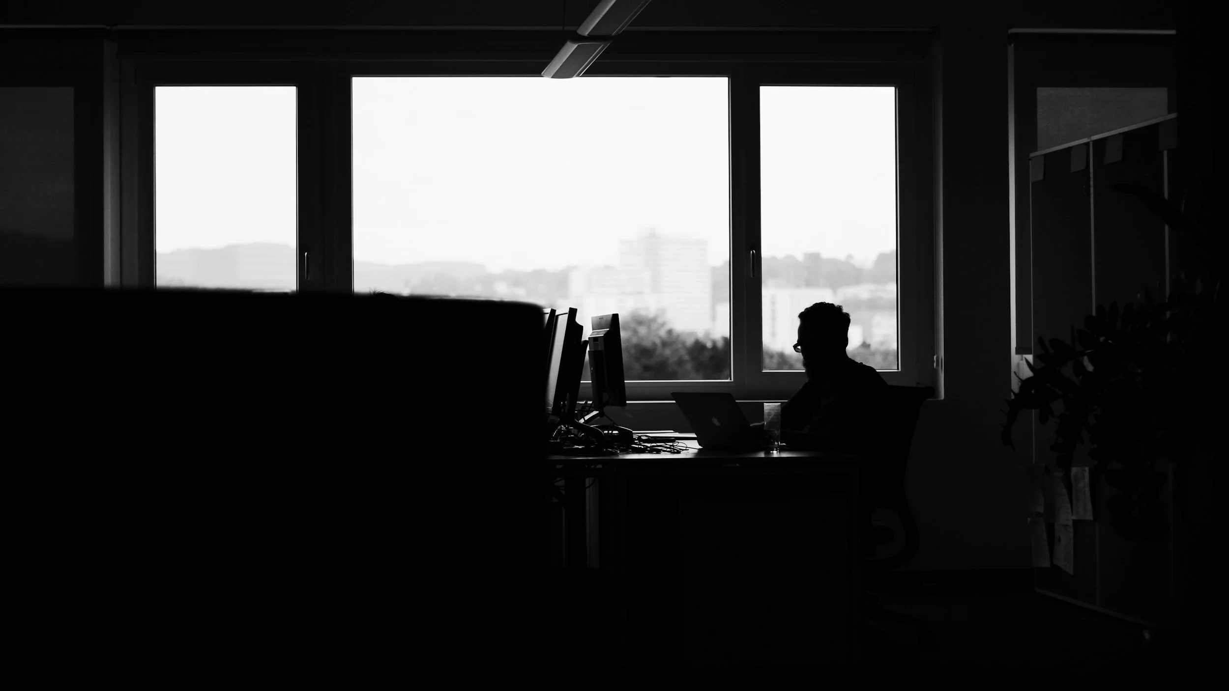 Silhouette of a person working at a desk in an office, with large windows showing a cityscape outside, in black and white.