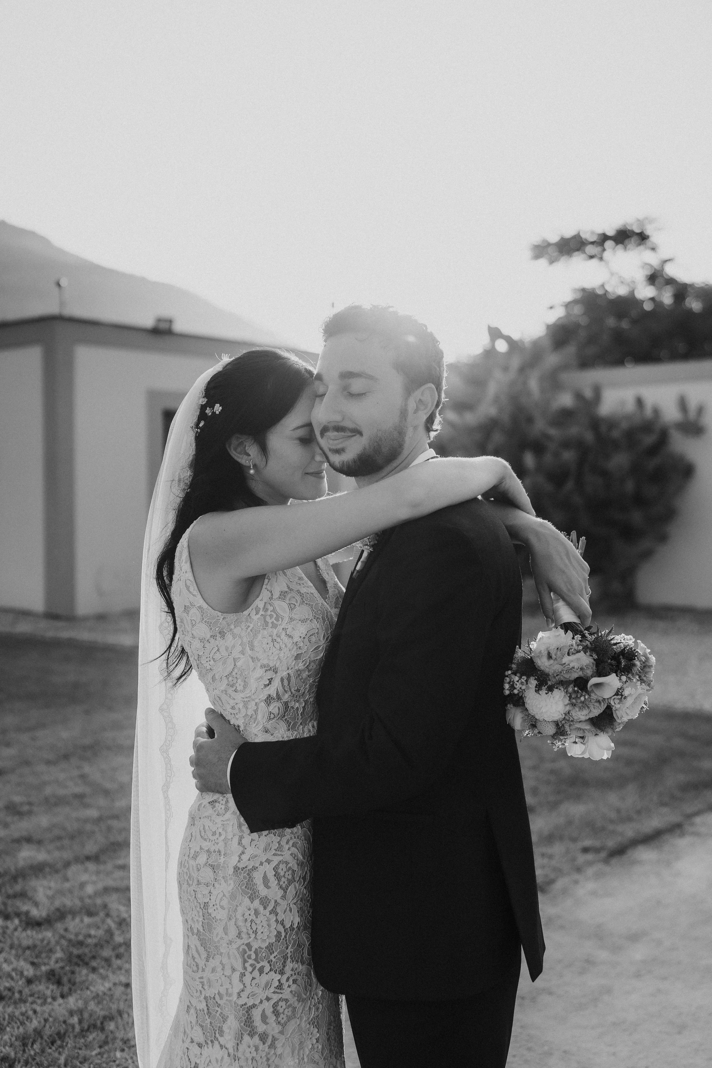 Due persone, una donna in abito da sposa e un uomo in abito, si abbracciano romantically all'aperto con un paesaggio di montagna e un edificio sullo sfondo.