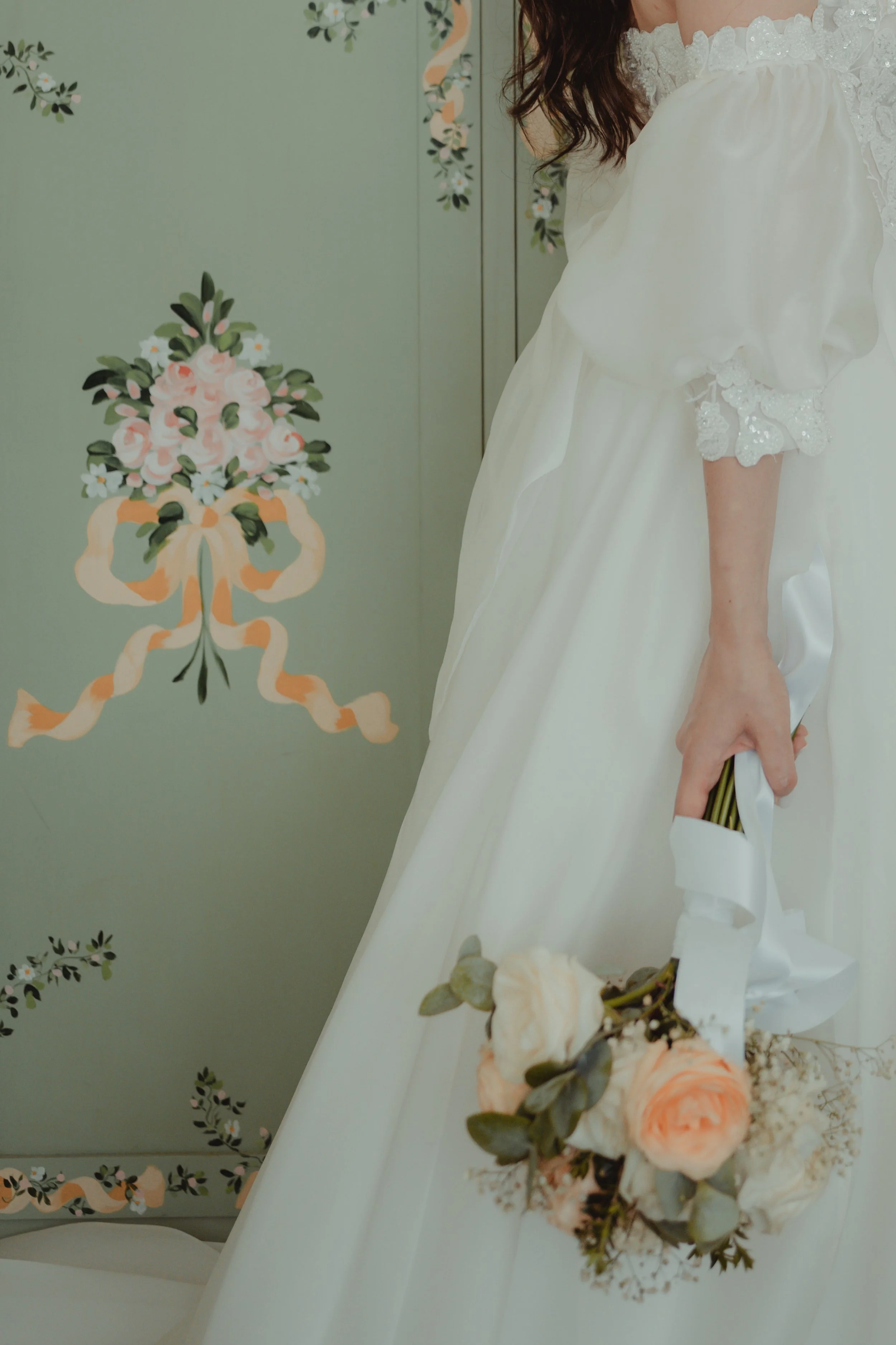 Immagine di una donna in abito da sposa con vaso di fiori sulla gonna, parte del volto e delle mani visibili, sfondo decorativo con motivi floreali.