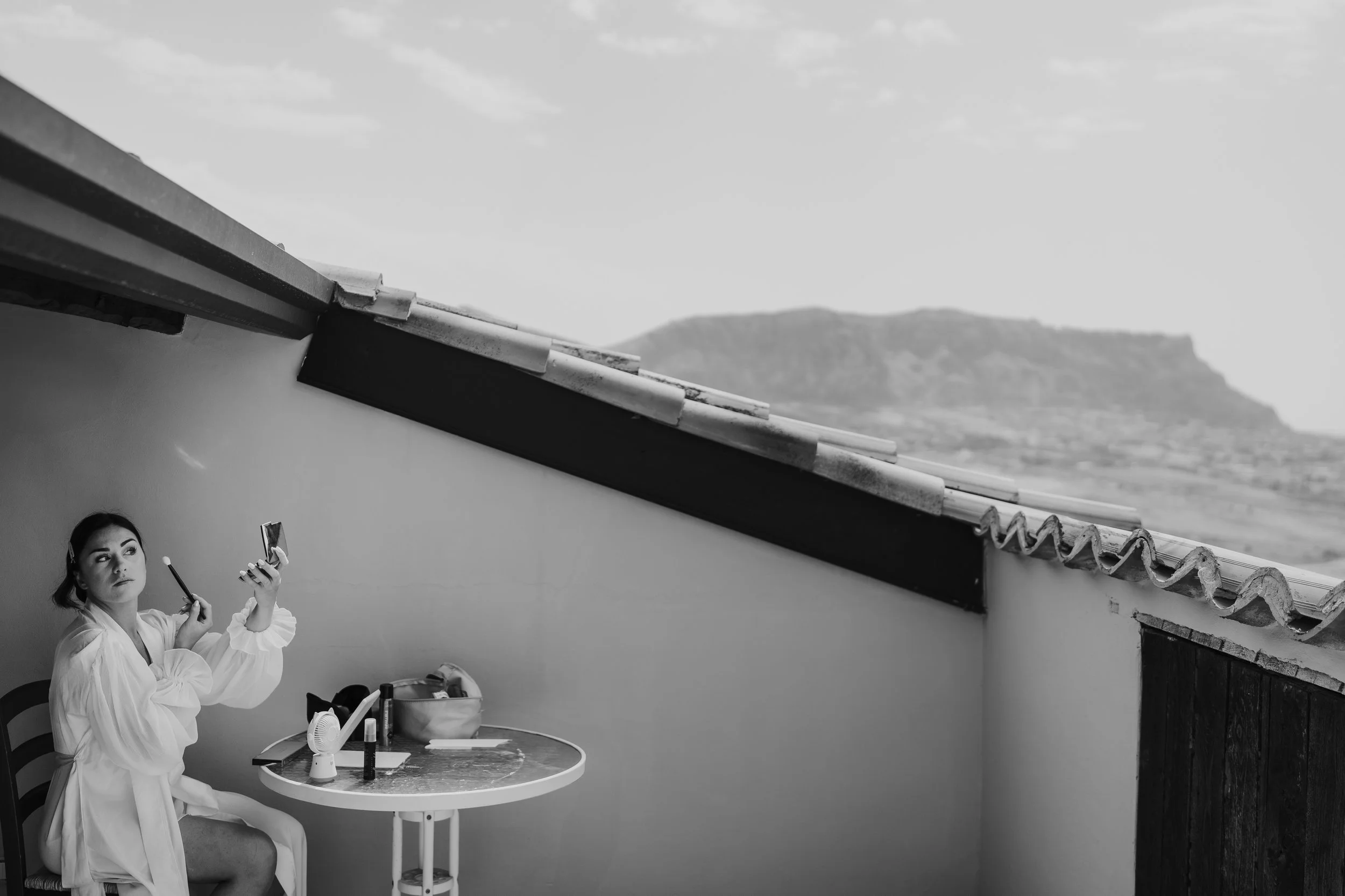 Donna in abito bianco che si trucca di fronte a uno specchio rotondo su un tavolo con vari oggetti, con una parete bianca e una finestra che mostra un paesaggio montano.