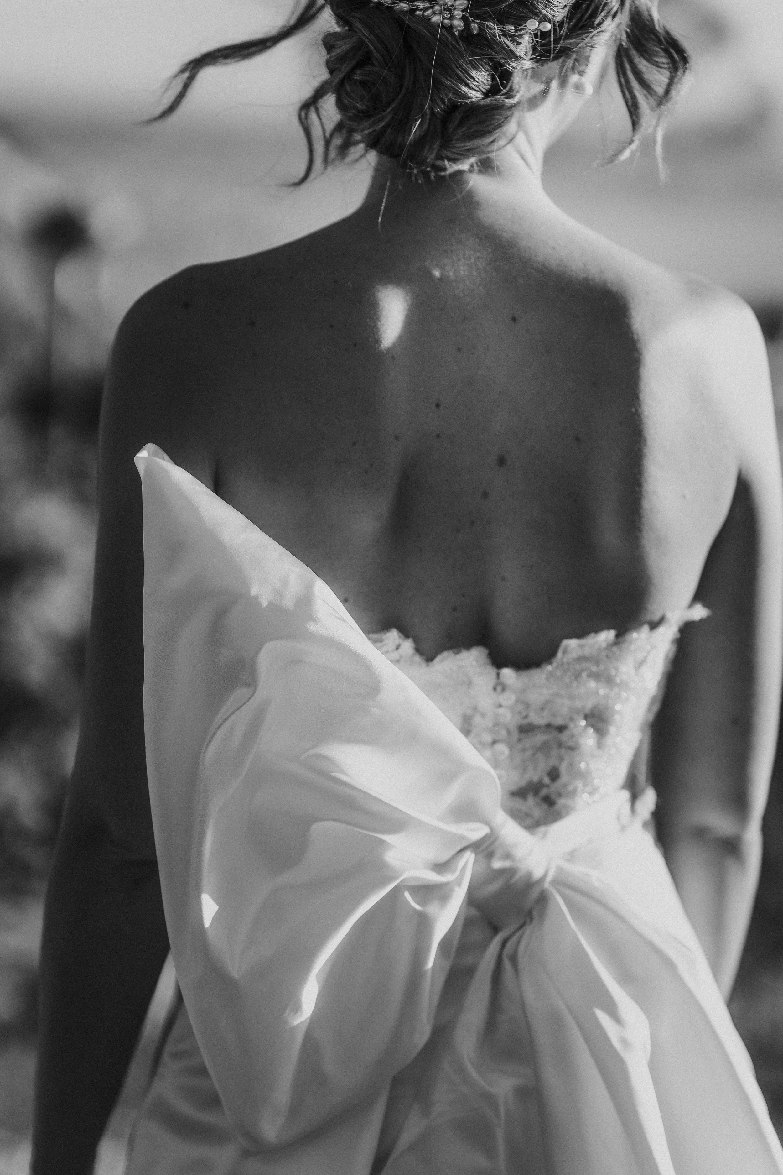Una donna con capelli corti e ricci, indossa un abito bianco da sposa e si trova sulla spiaggia, vista da dietro in una foto in bianco e nero.