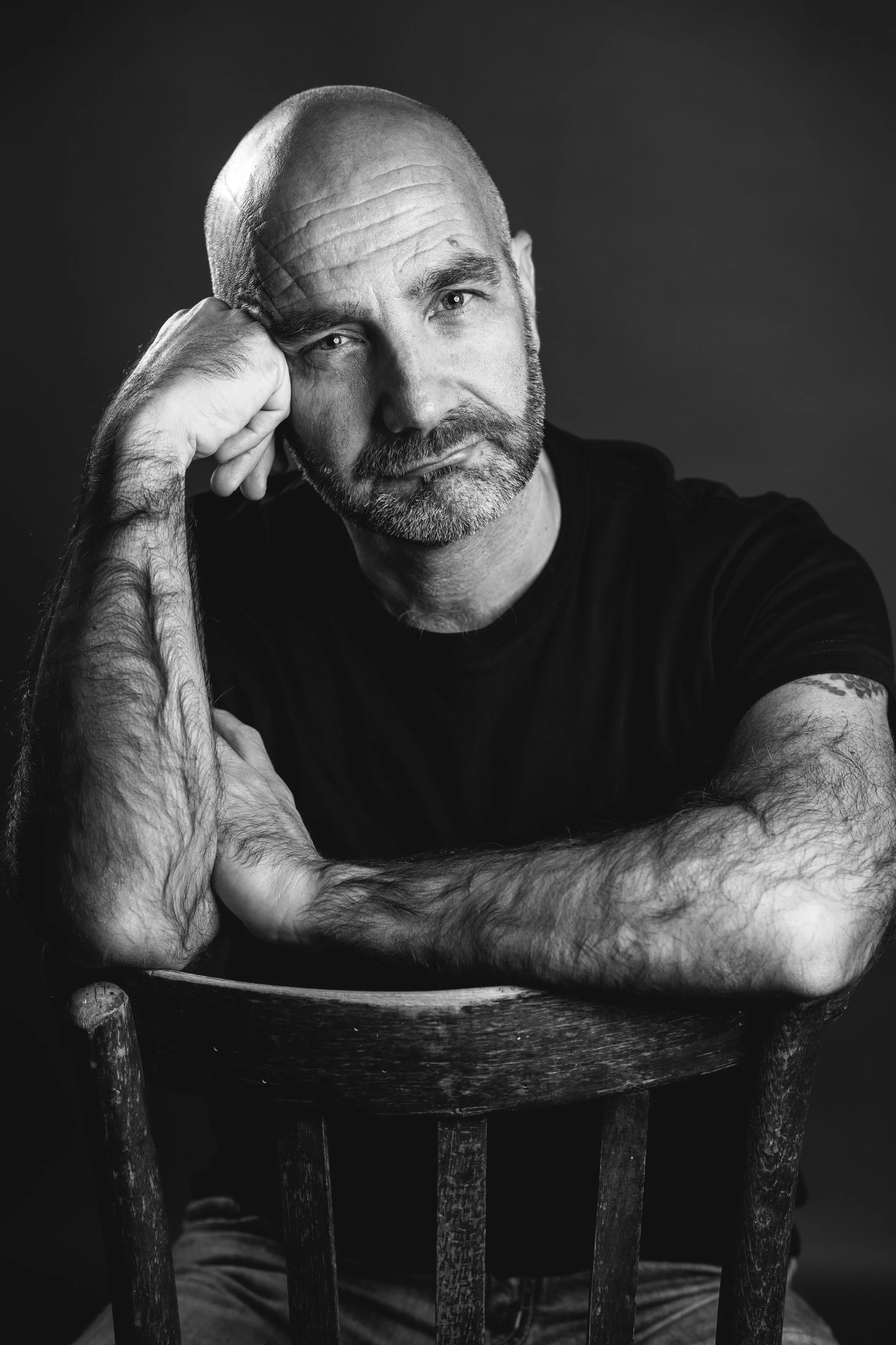 Ritratto in bianco e nero di un uomo con barba rasata, braccia incrociate su una sedia di legno, in posa relax, con il ginocchio visibile in basso.